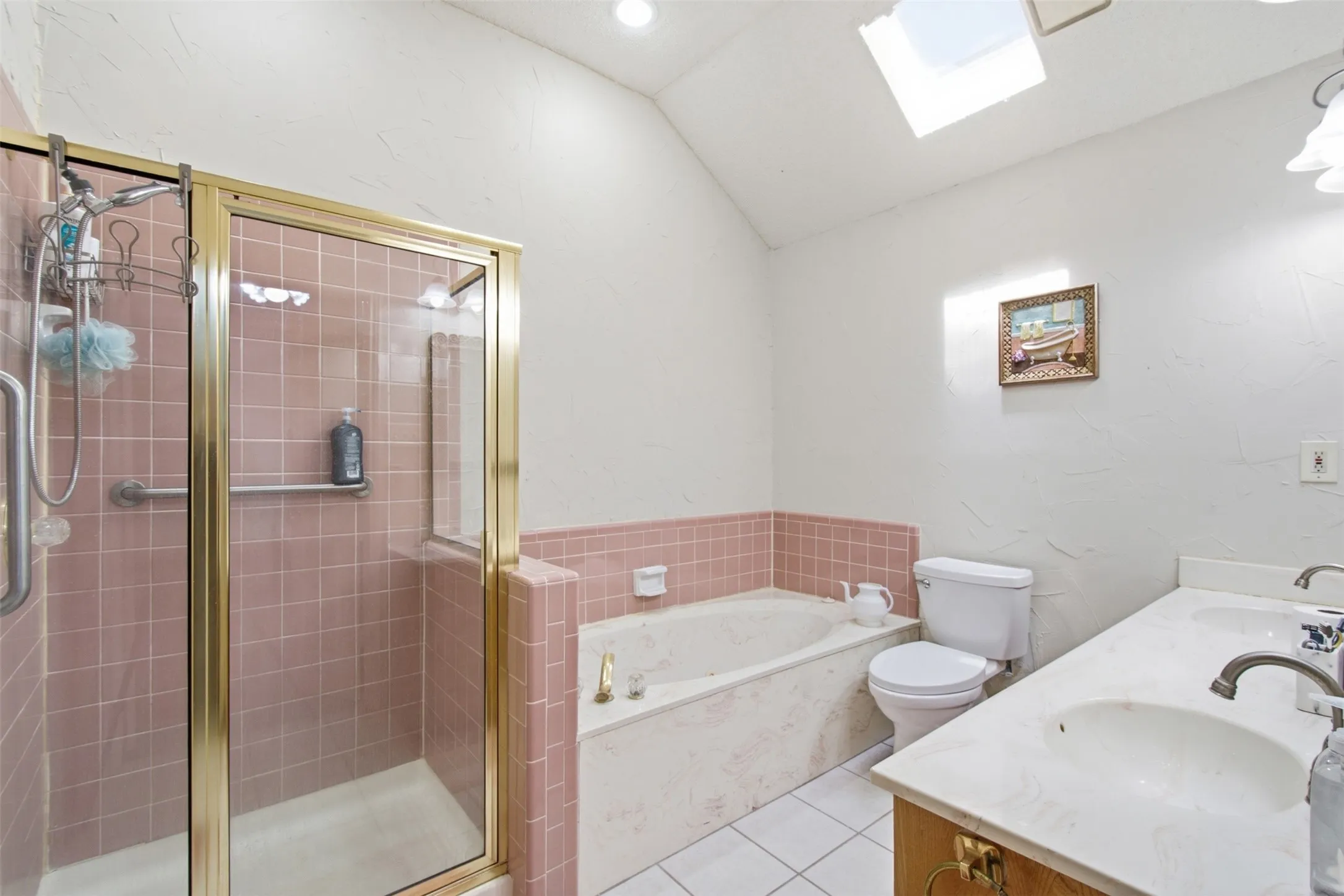Bathroom featuring a shower with gold-tone framed glass enclosure, an integrated bathtub, a dual-sink vanity with white countertops, and a skylight