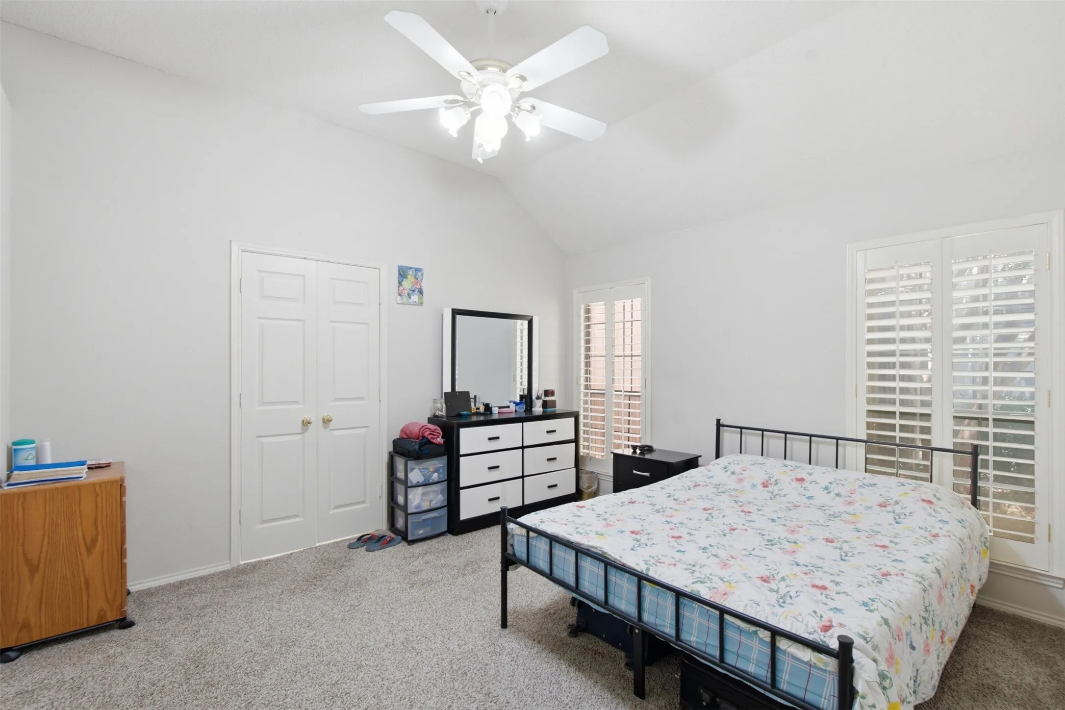 Vaulted ceiling with a ceiling fan, white paneled double doors, two windows with white plantation shutters, and gray carpeting