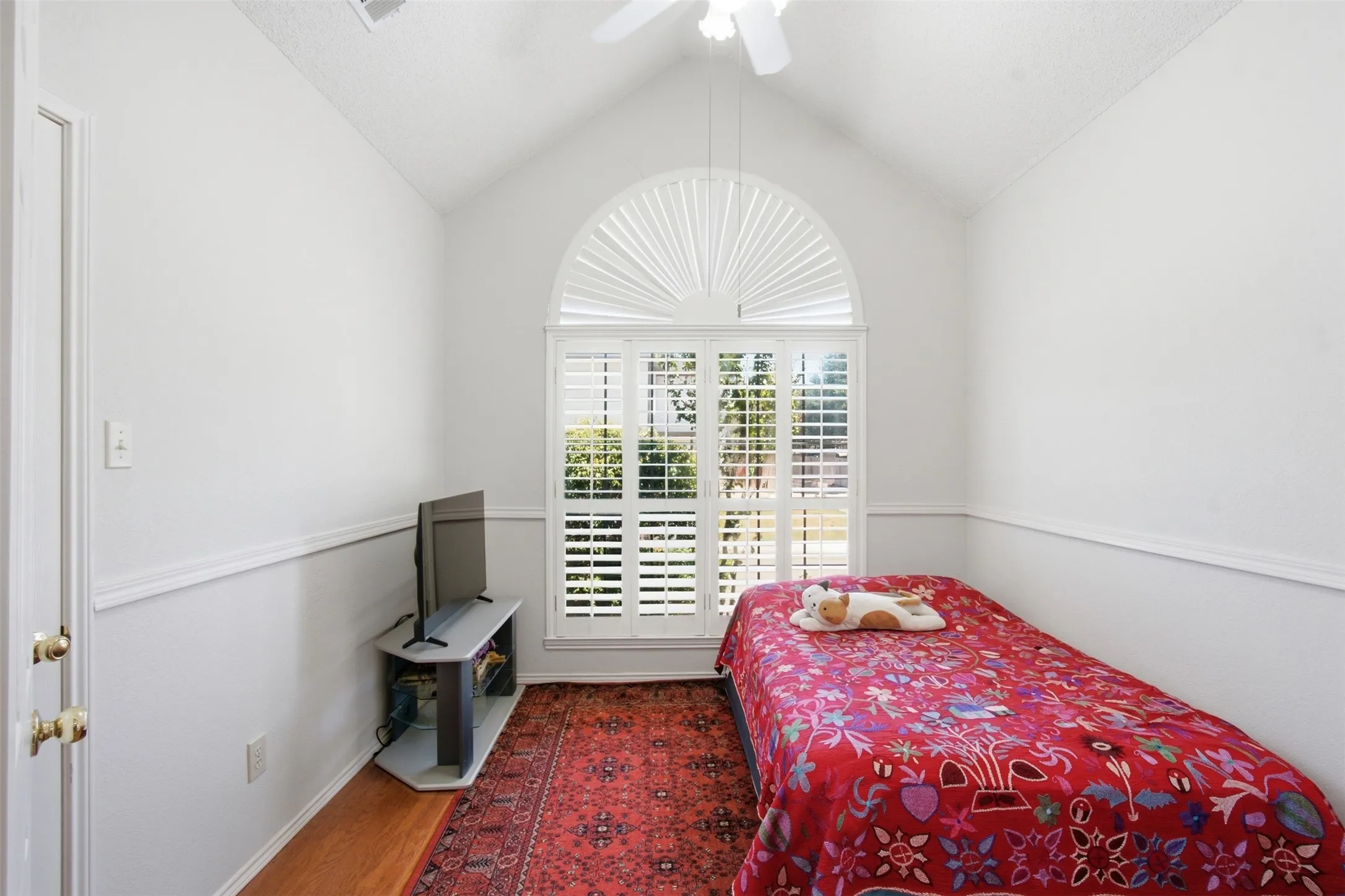 Vaulted ceiling featuring a ceiling fan, arched window with plantation shutters, wood-finish flooring, and chair rail molding