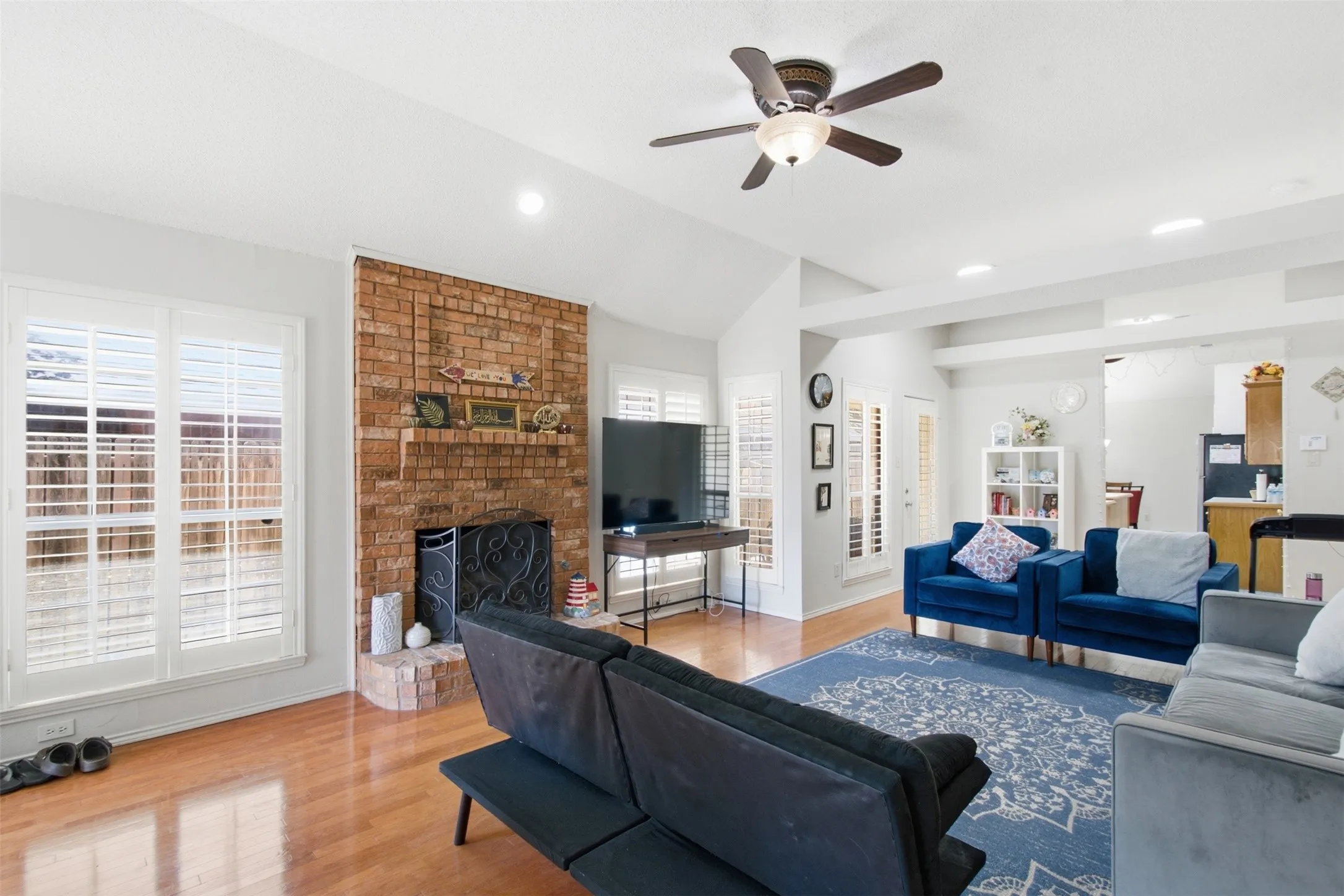 Brick-faced fireplace with a hearth, wood-finish flooring, white window shutters, vaulted ceiling, and a ceiling fan
