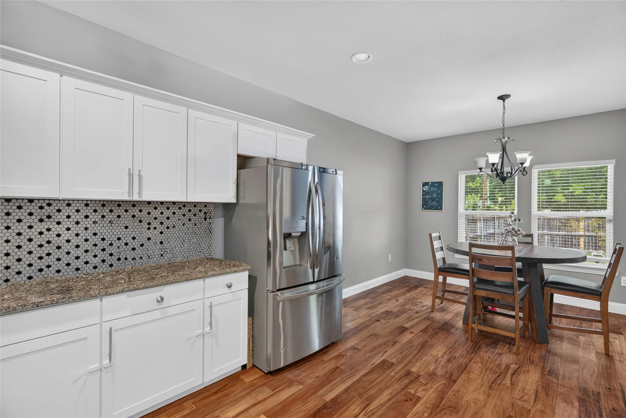 White shaker cabinetry, honeycomb tile backsplash, stainless steel refrigerator, wood-finish flooring, and a multi-light chandelier