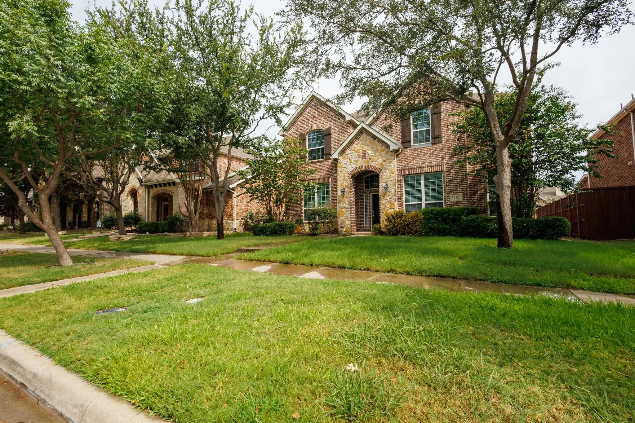 Brick and stone facade residence with mature trees, a manicured lawn, and defined shrubbery