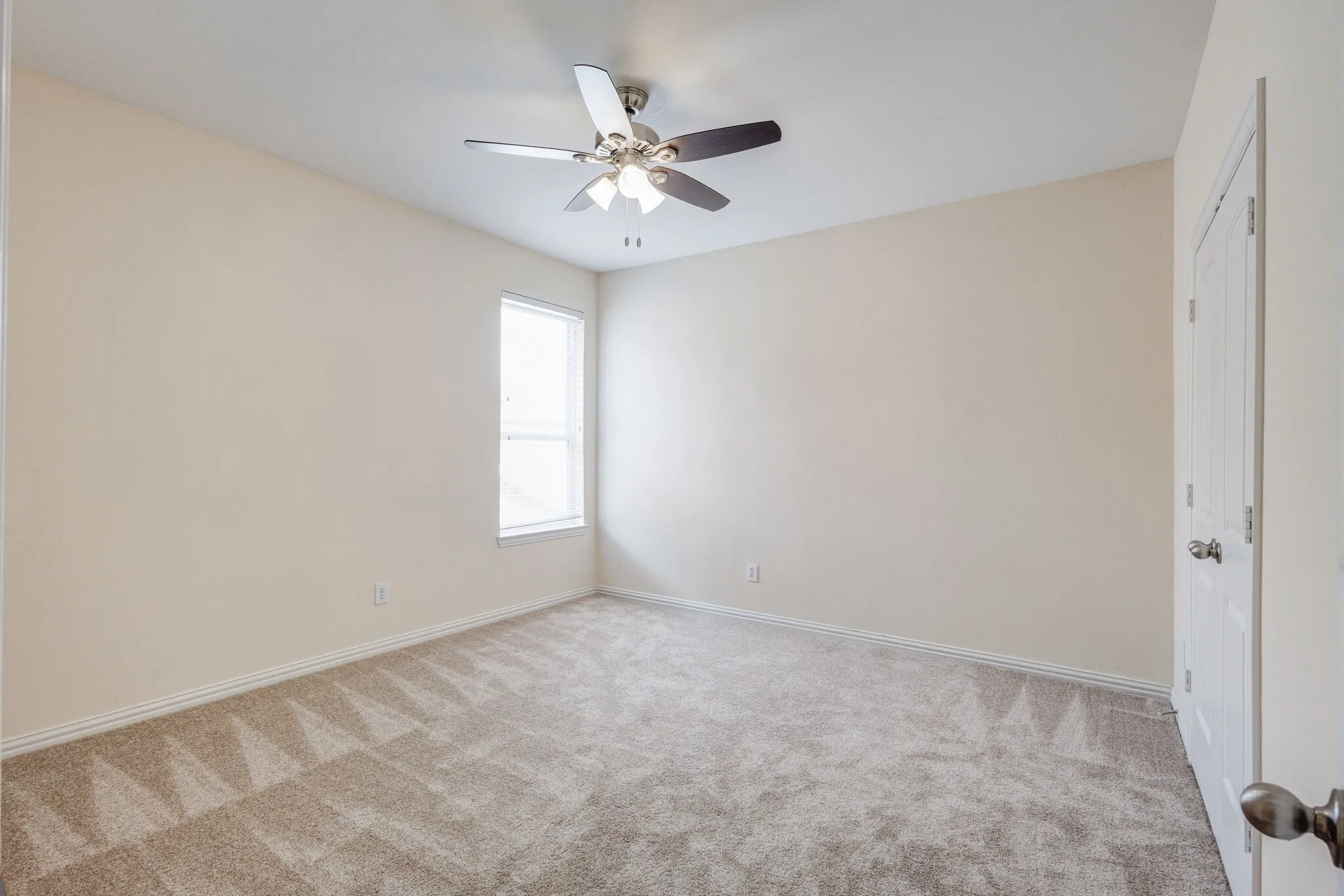 Neutral-toned walls, plush carpet flooring, a window with white trim, a ceiling fan with integrated lighting, and a white interior door with a silver-tone knob