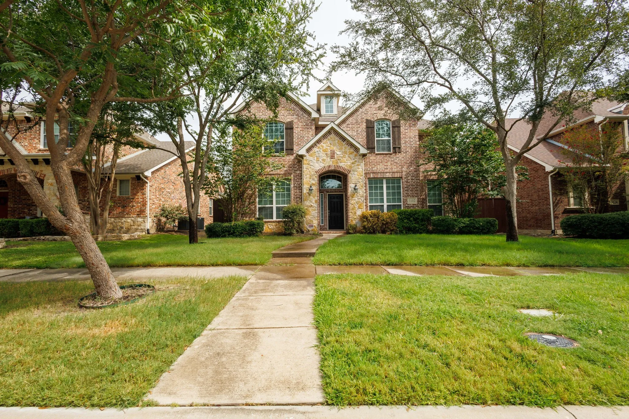 Brick and stone facade, dormer window, established trees, concrete walkway, and manicured lawn
