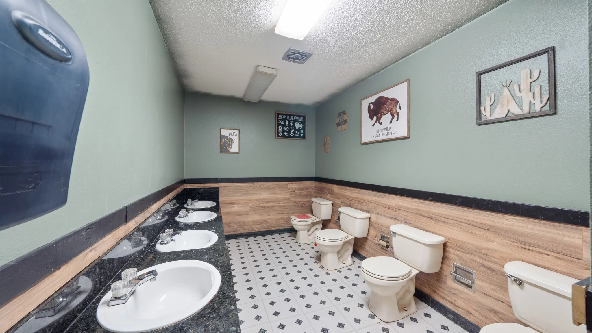 Multiple vanity sinks with dark -countertops, light green walls, wood-finish wainscoting, and patterned floor tiles