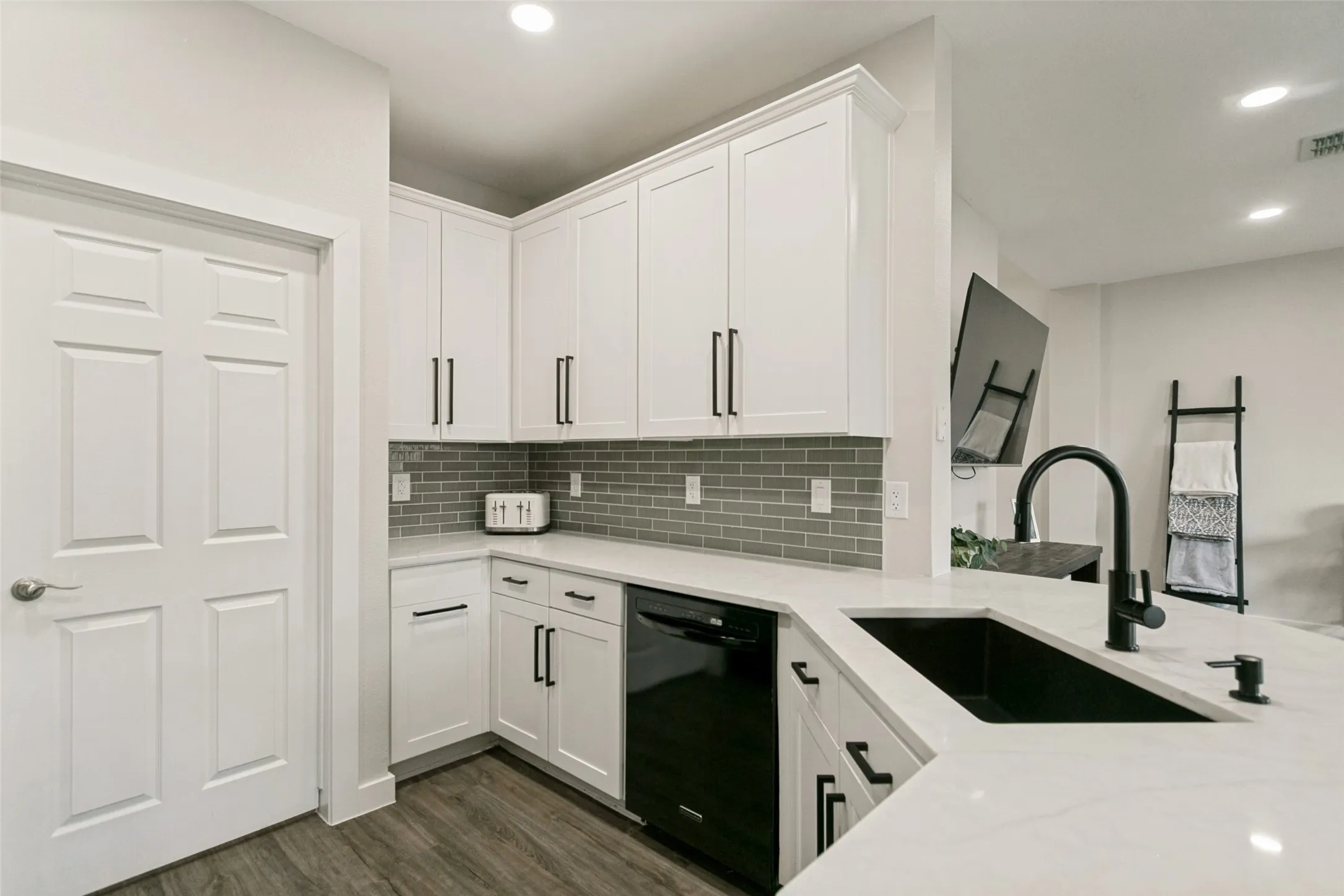 White cabinetry, light countertop surfaces, a dark-finish kitchen sink, a tiled backsplash, and wood-finish flooring