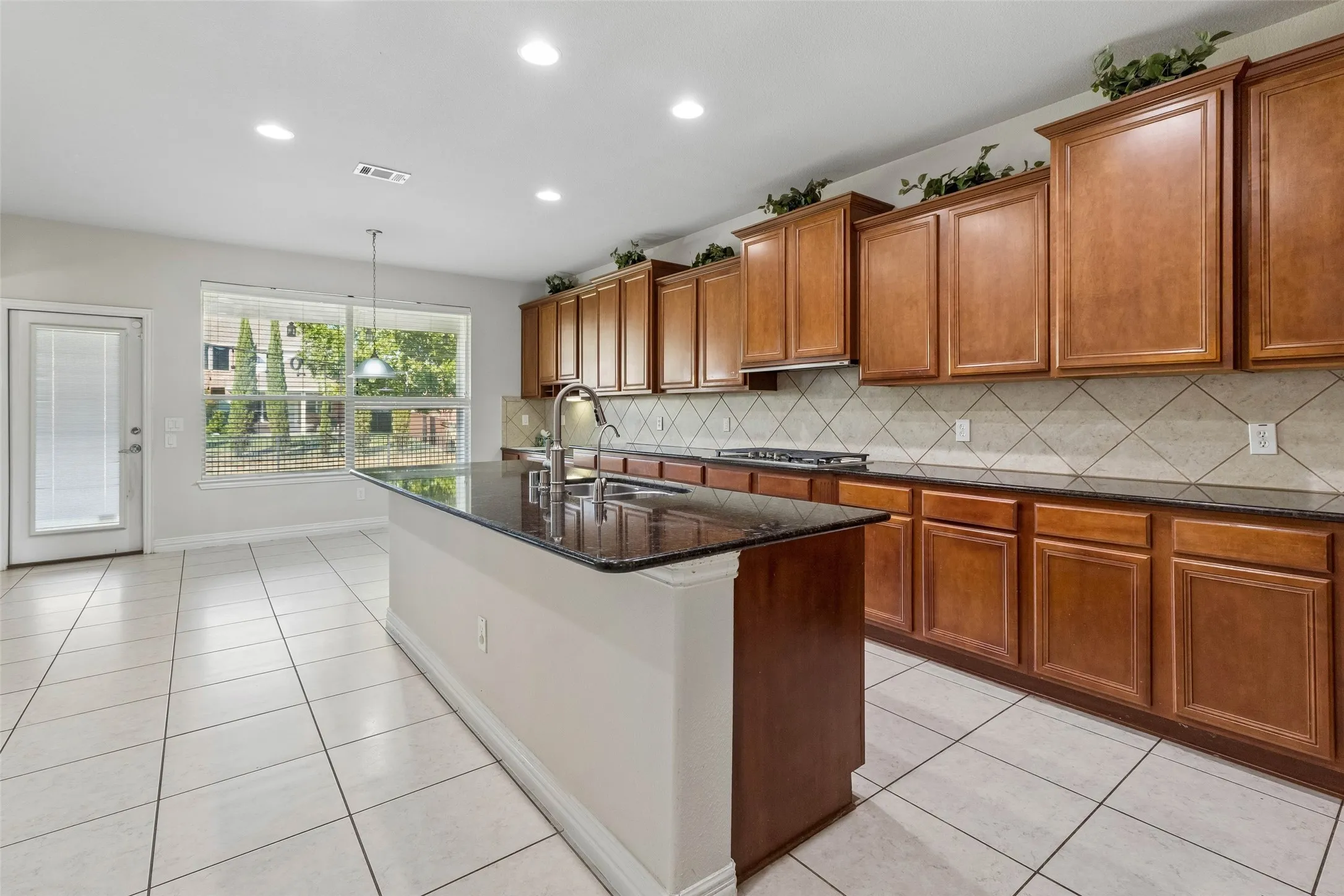 Kitchen featuring an island with an integrated sink, dark countertops, wood cabinetry, a tiled backsplash, and recessed lighting