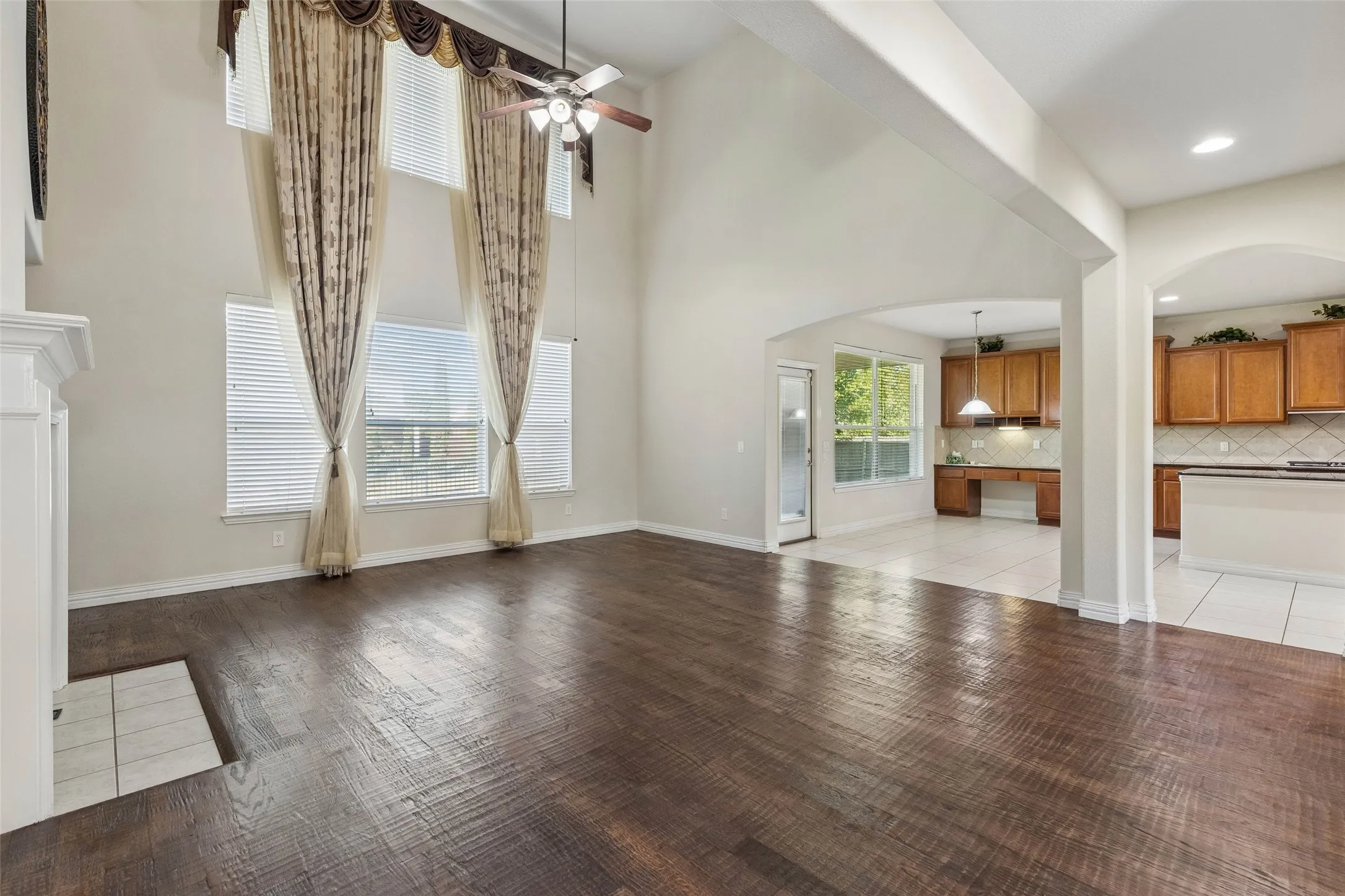 Vaulted ceiling with a ceiling fan, wood-finish flooring, white fireplace surround, multiple tall windows with blinds, and an arched entryway to the kitchen