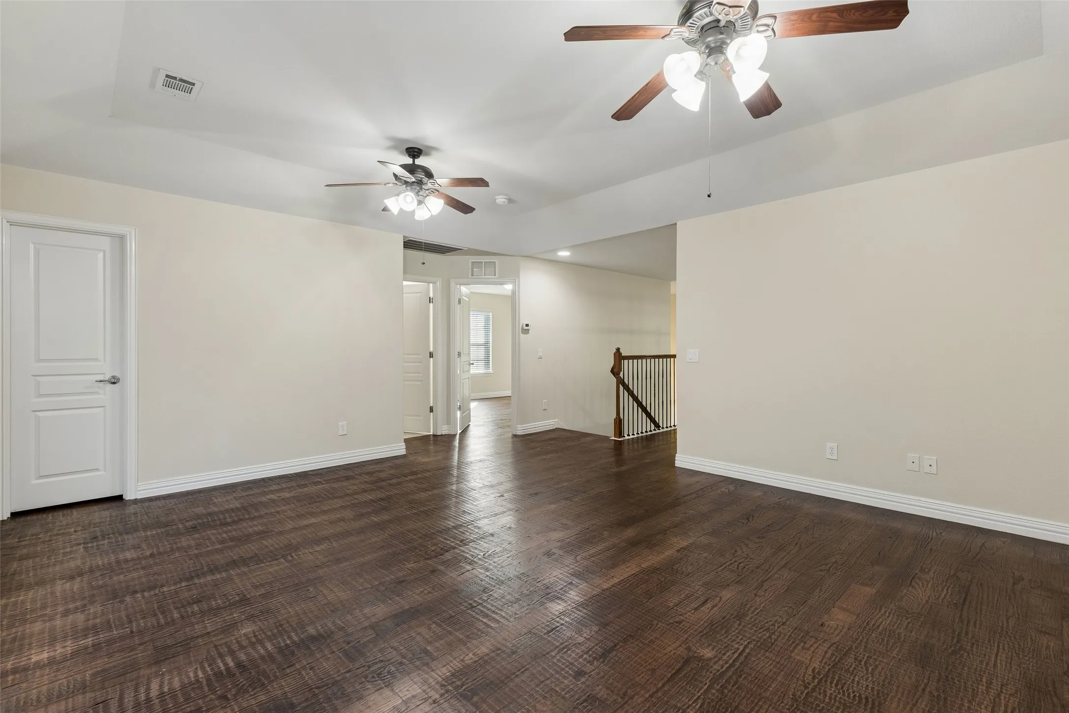 Expansive room featuring wood-finish flooring, tray ceiling, two ceiling fans, and staircase with wooden handrail