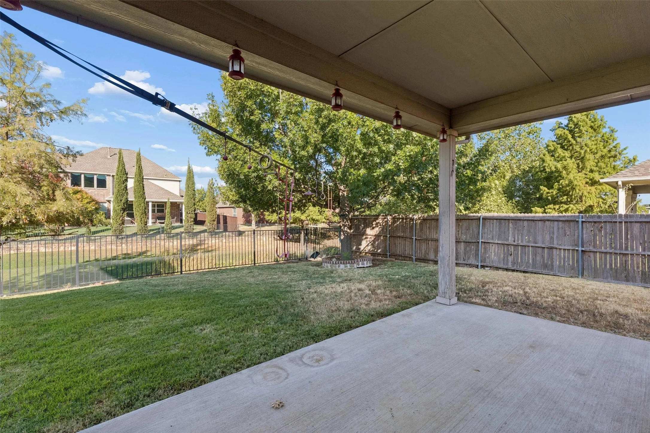 Covered concrete patio with overhead lighting, overlooking a grass yard with a metal fence and mature trees
