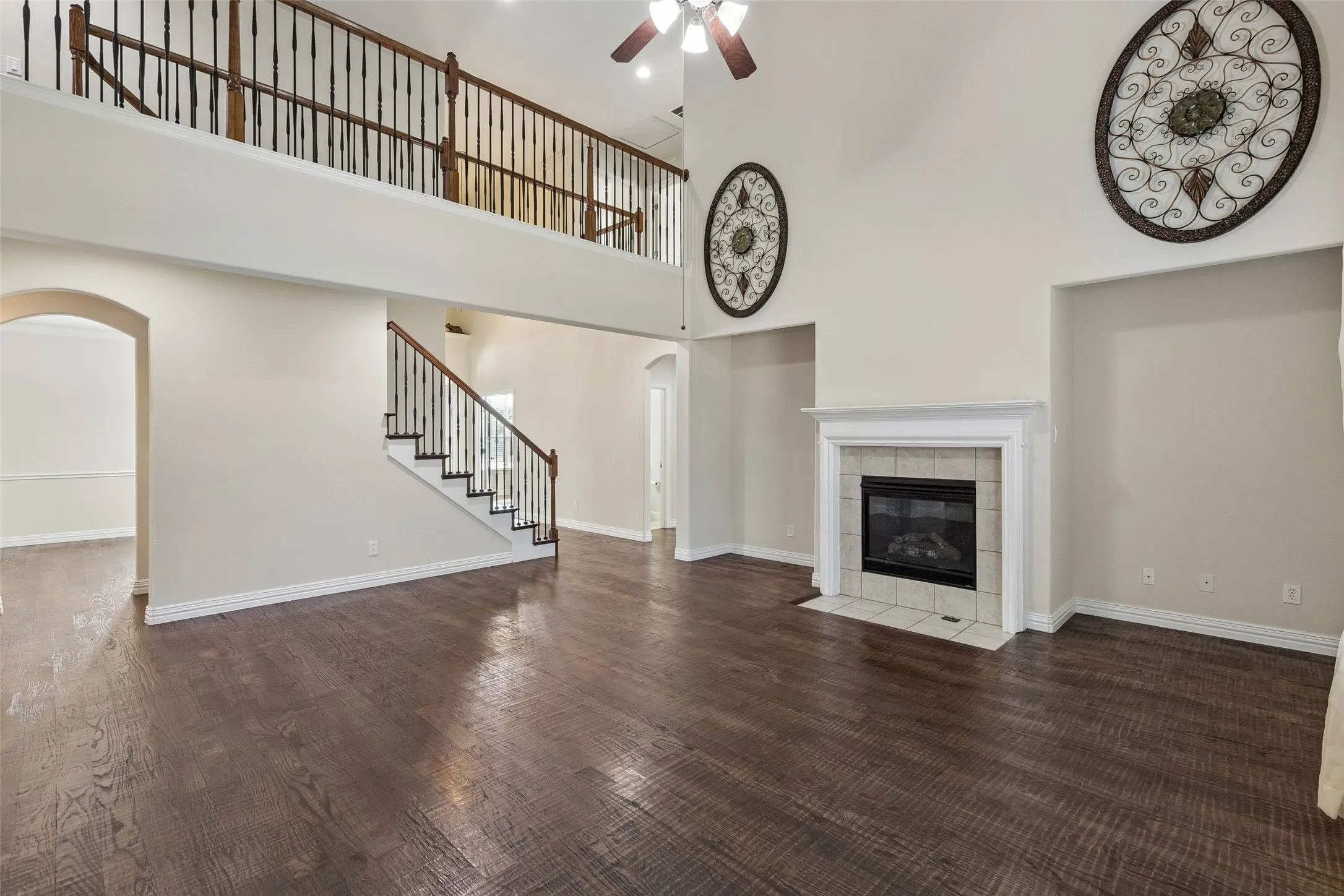 Expansive living area featuring high ceilings, dark wood-finish flooring, a tiled fireplace with a white mantel, an arched doorway, and a second-story landing with wood railings