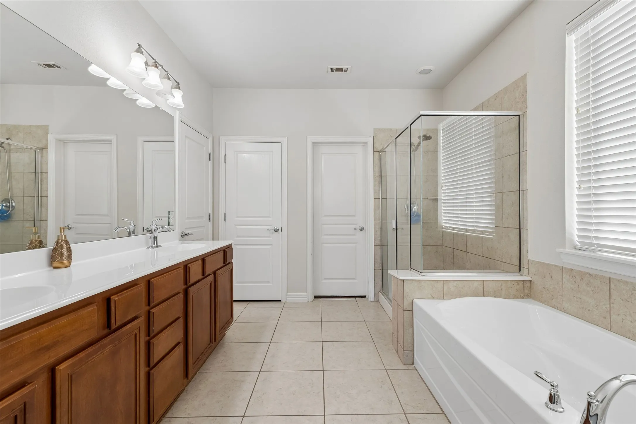 Dual vanity with wood-finish cabinetry and white countertops, large mirror, walk-in shower with glass enclosure, white soaking tub, and tiled flooring