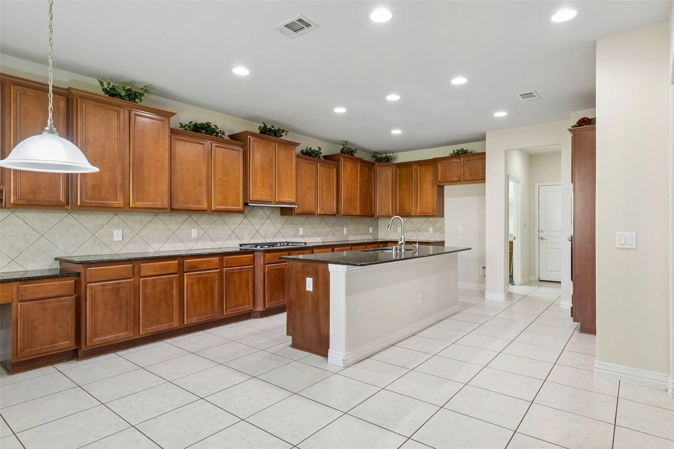 Spacious kitchen featuring extensive wood cabinetry, dark countertops, a tiled backsplash, a central island with a sink, and recessed lighting