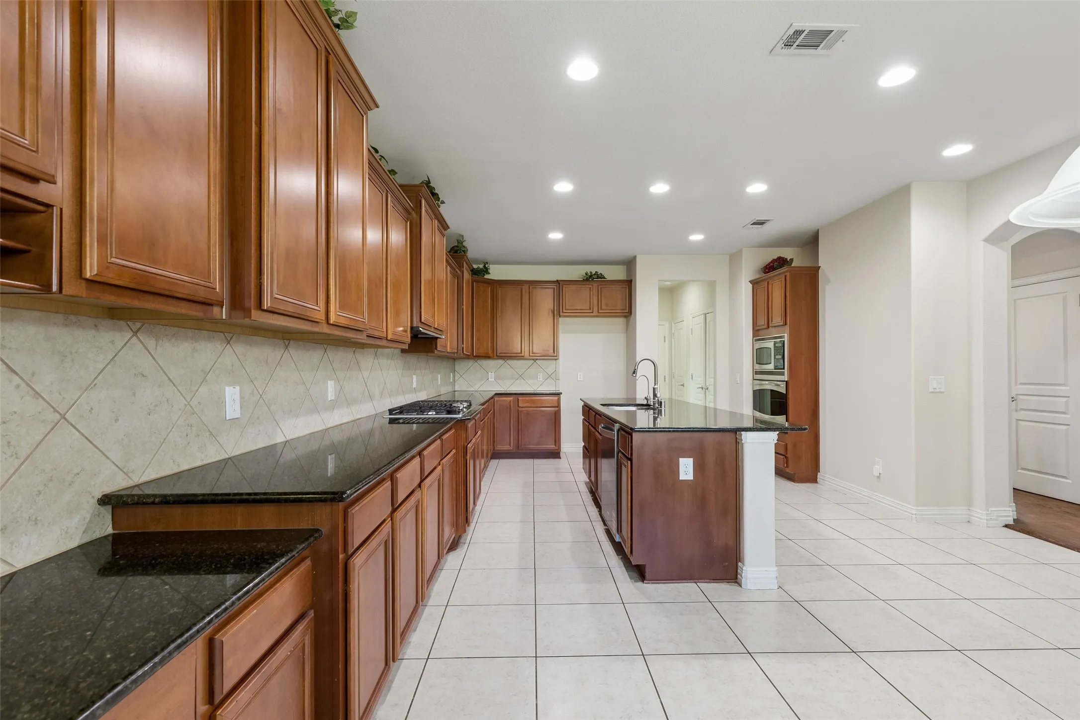 Kitchen featuring wood cabinetry, dark countertops, a center island with sink, tile flooring, and recessed lighting