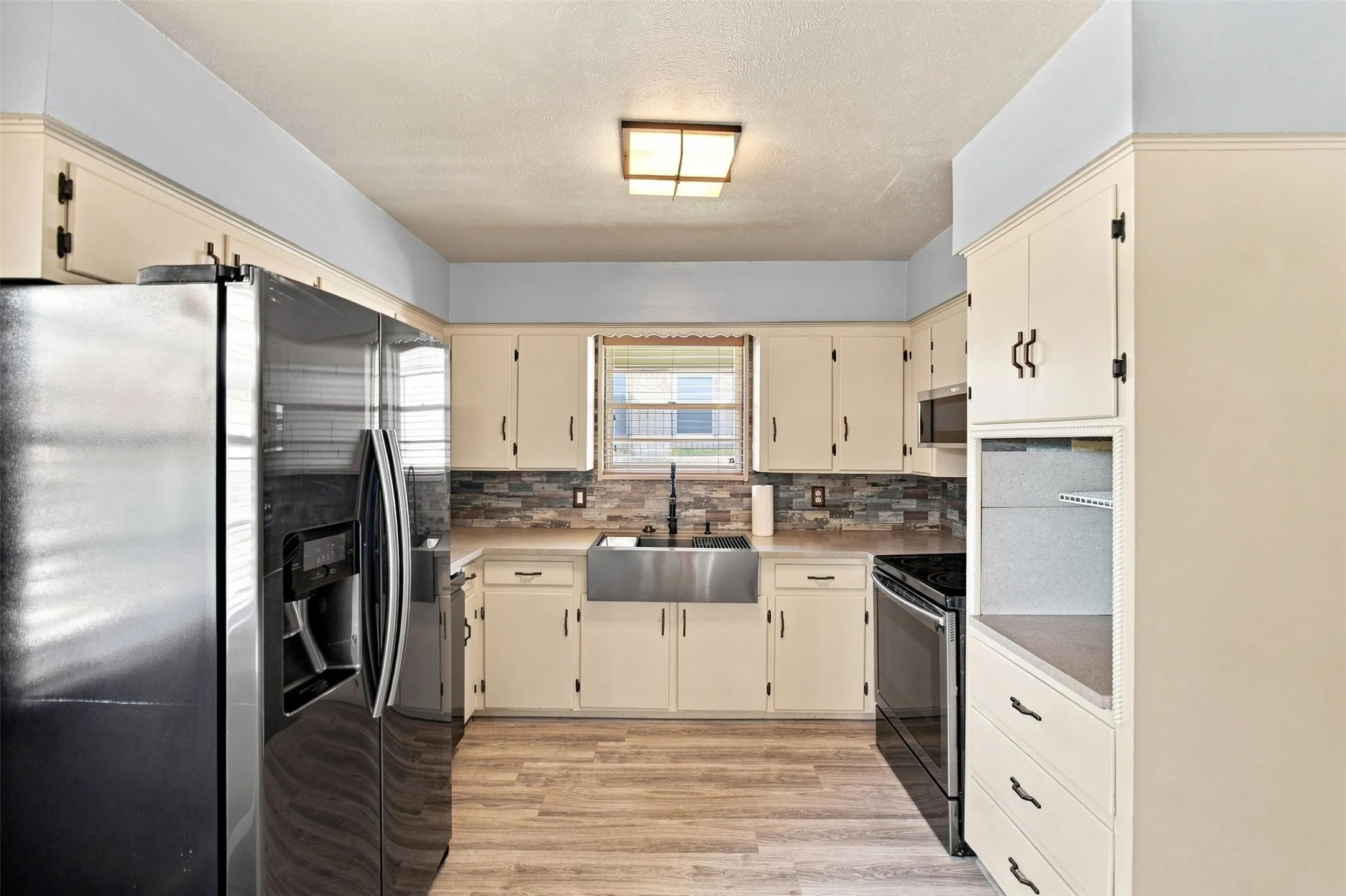 Kitchen featuring wood-finish flooring, a stainless steel farmhouse sink, stacked stone backsplash, and cream cabinetry with dark hardware