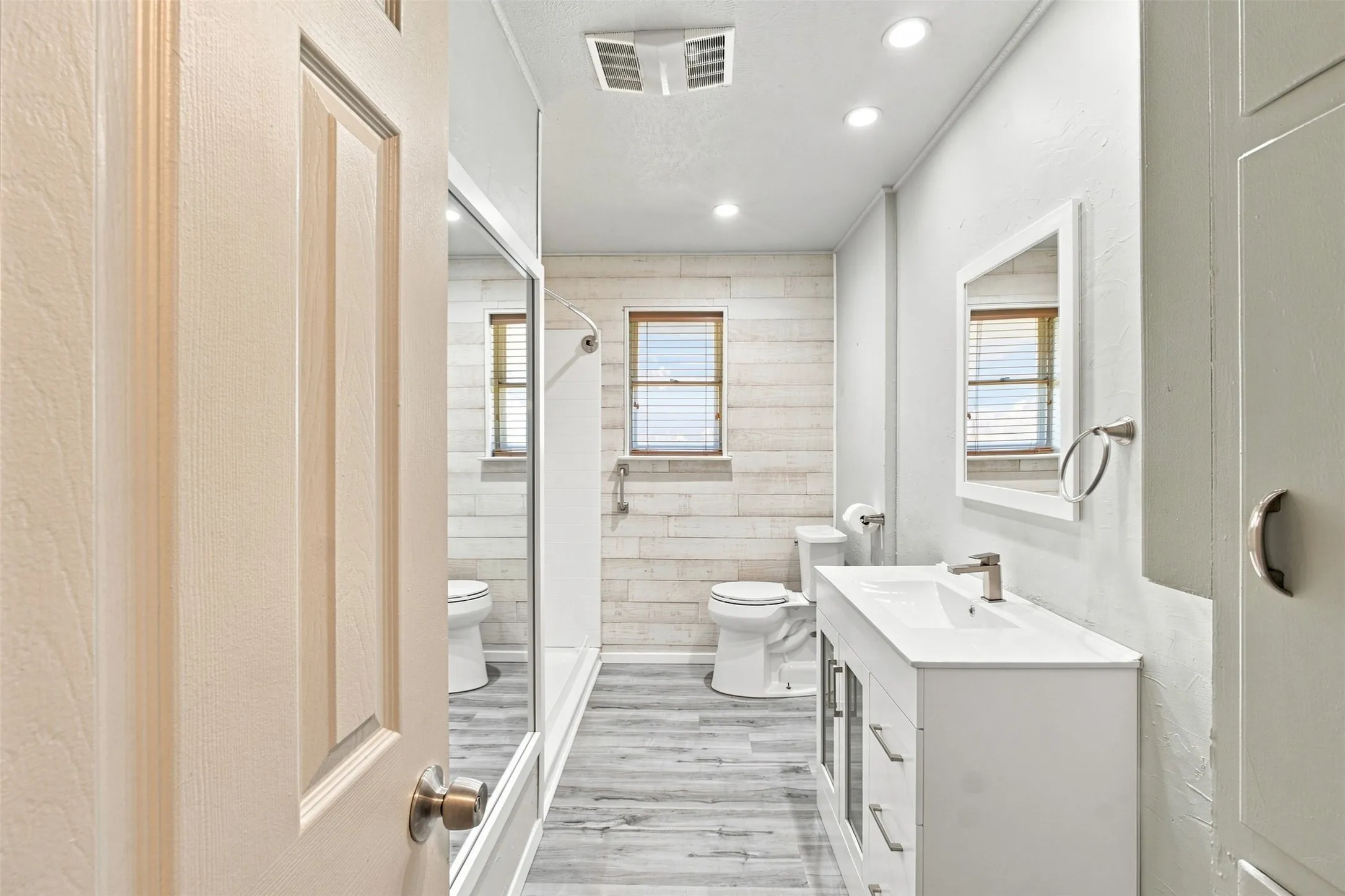 Modern bathroom featuring wood-finish flooring, a white vanity with integrated sink, recessed lighting, and a walk-in shower with glass enclosure