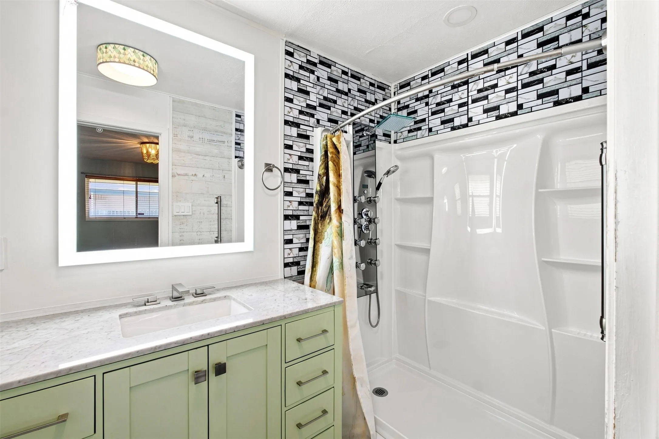 Illuminated vanity mirror, light green custom cabinetry, white countertop with integrated sink, black and white mosaic tile shower surround, and white shower enclosure