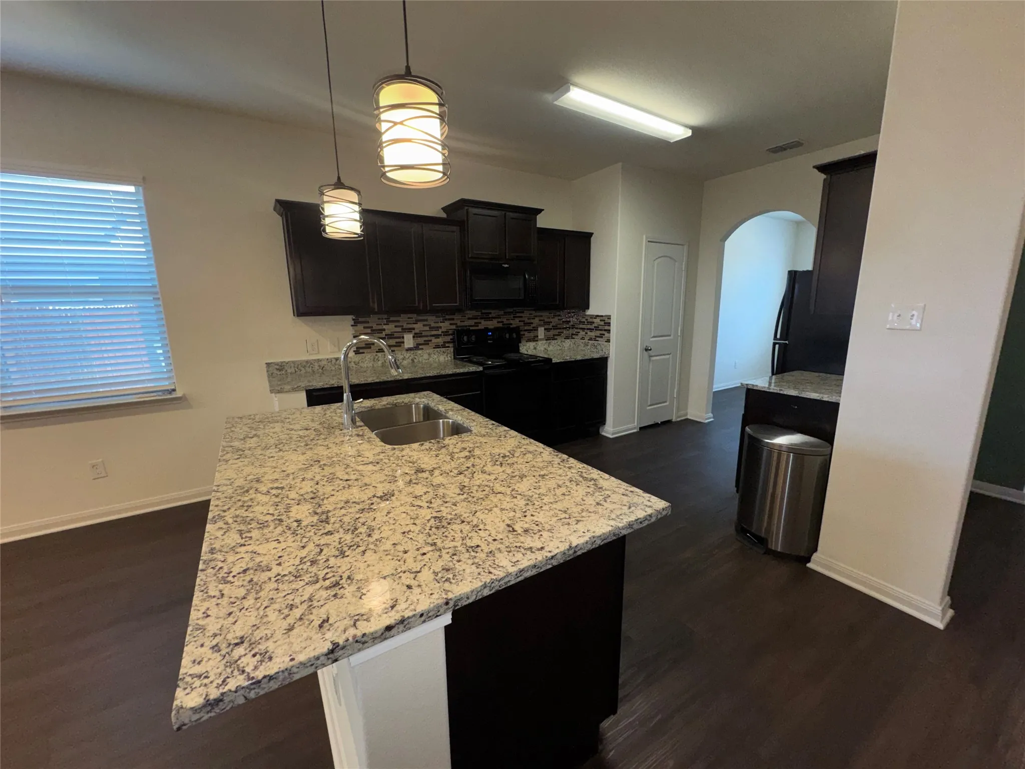 Kitchen featuring an island with a double basin sink, dark wood-finish cabinetry, light-toned countertops, tile backsplash, and dark wood-finish flooring
