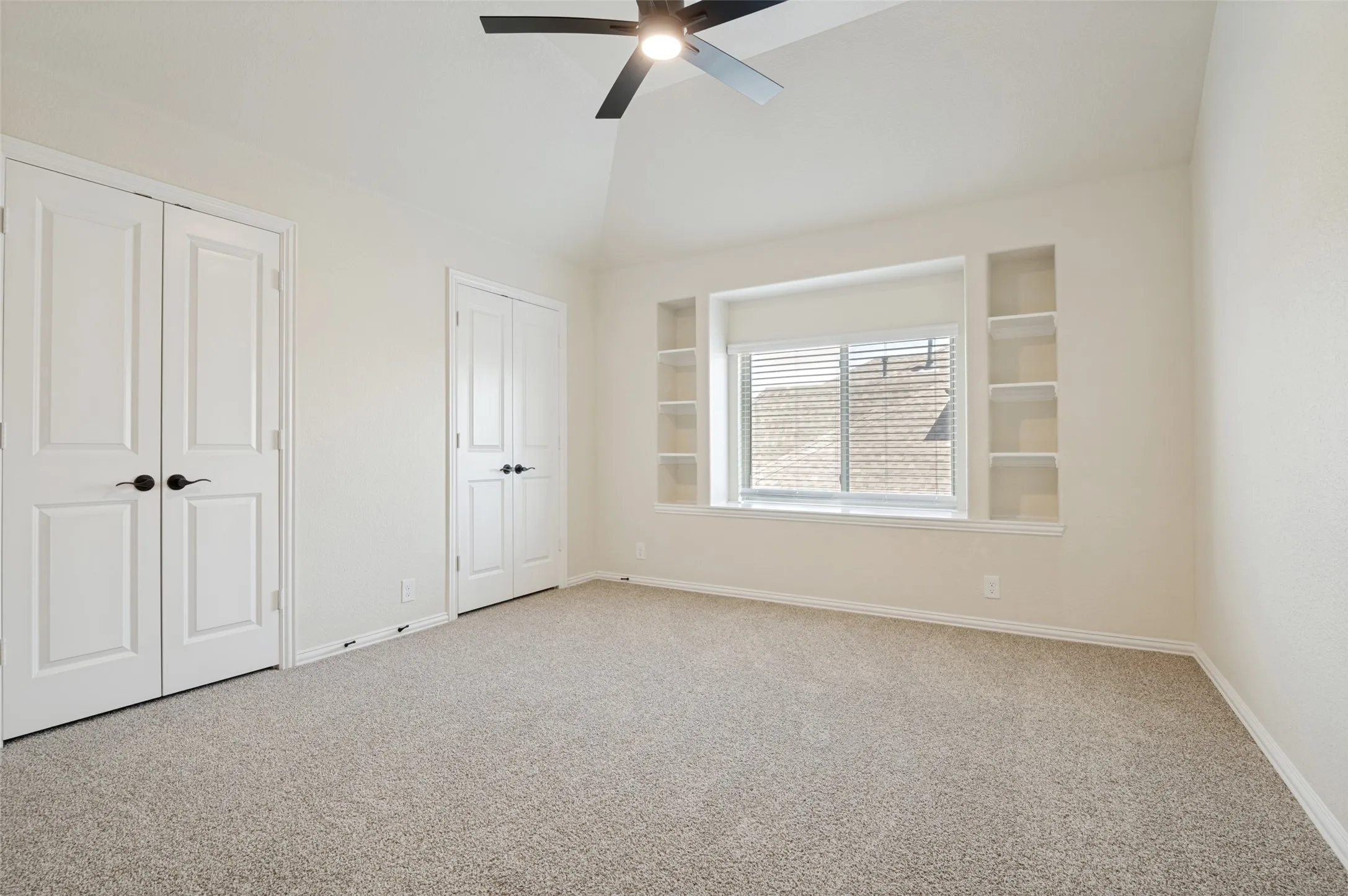 Vaulted ceiling with a ceiling fan, neutral carpet, built-in shelving, two sets of double doors, and a window with blinds
