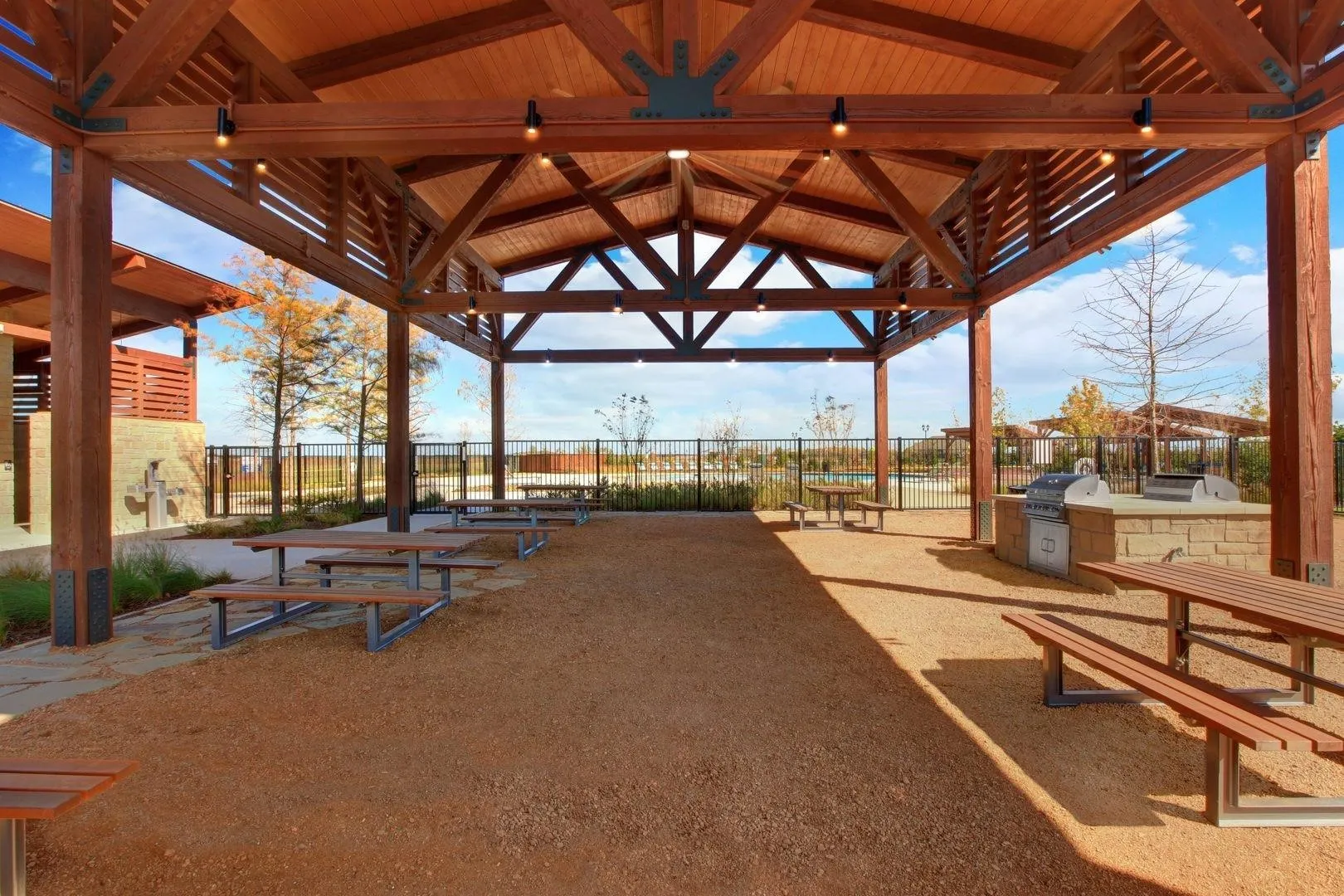 Expansive covered outdoor structure featuring a substantial wood beam roof, built-in BBQ grills, multiple picnic tables, and a gravel surface