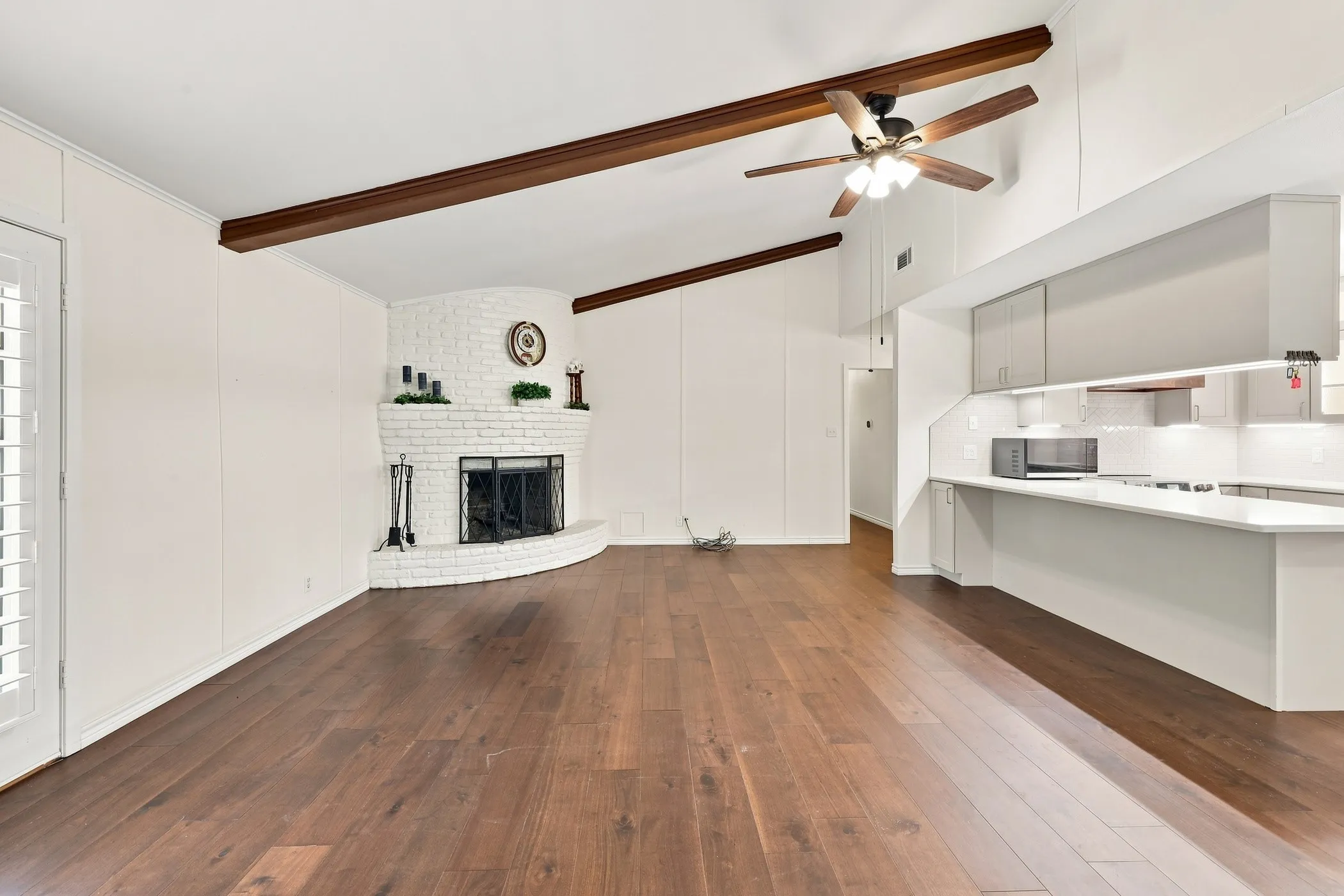 Vaulted ceiling with exposed wood beams, wood-finish flooring, white brick fireplace, modern kitchen cabinetry, and recessed lighting