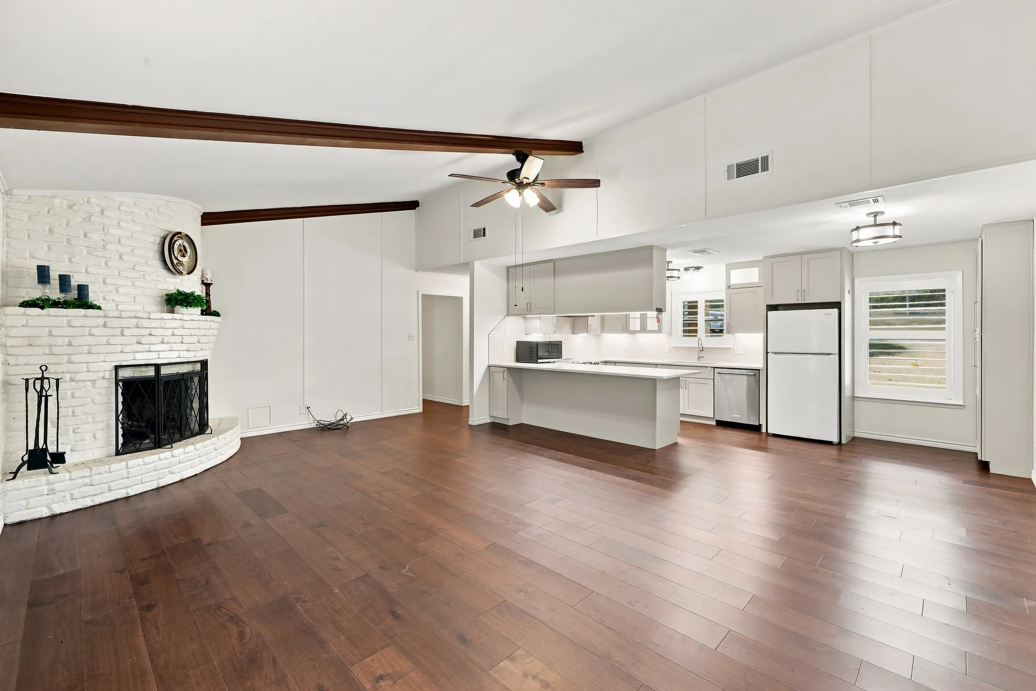 Open-concept living area featuring wood-finish flooring, a white-painted brick fireplace, an exposed beam ceiling, a ceiling fan, and recessed lighting in the kitchen area