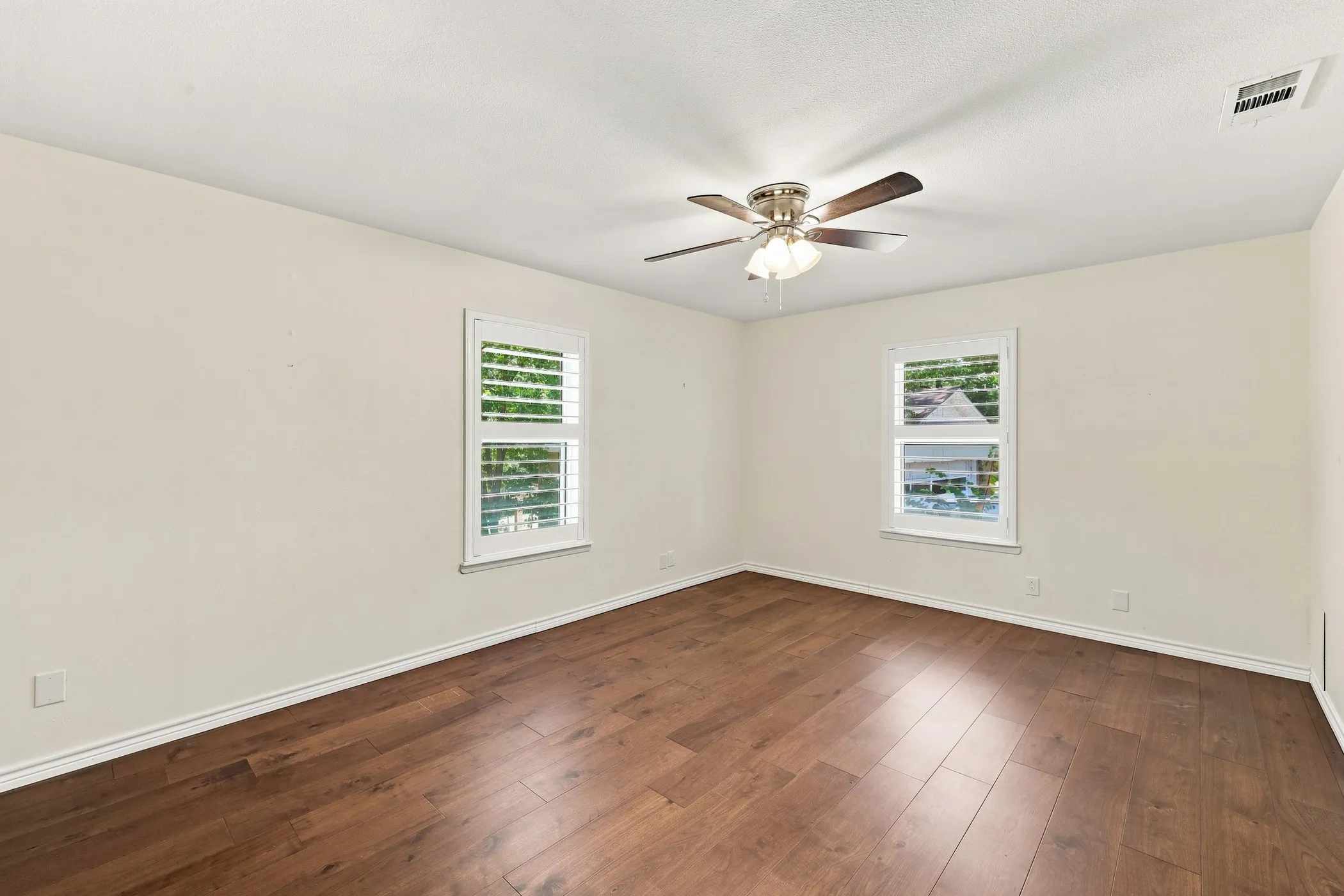 Wood-finish flooring, two windows with plantation shutters, ceiling fan with integrated lighting, and neutral wall tones
