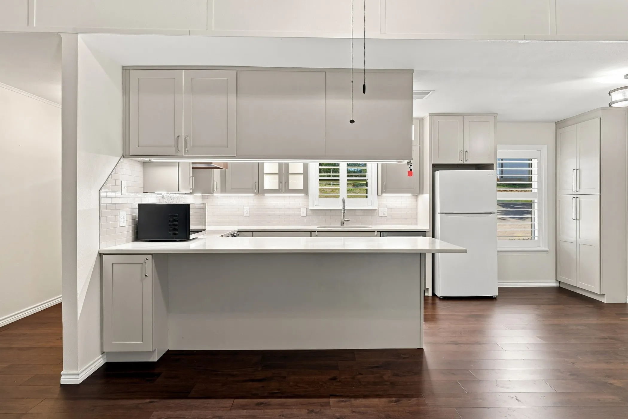 Contemporary kitchen featuring light gray shaker-style cabinetry, a white subway tile backsplash, a peninsula with a white countertop, and dark wood-finish flooring