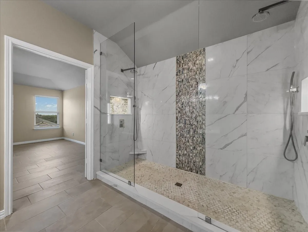 Walk-in shower with clear glass enclosure, marble-patterned tile, mosaic tile accent, rainfall showerhead, and pebble flooring