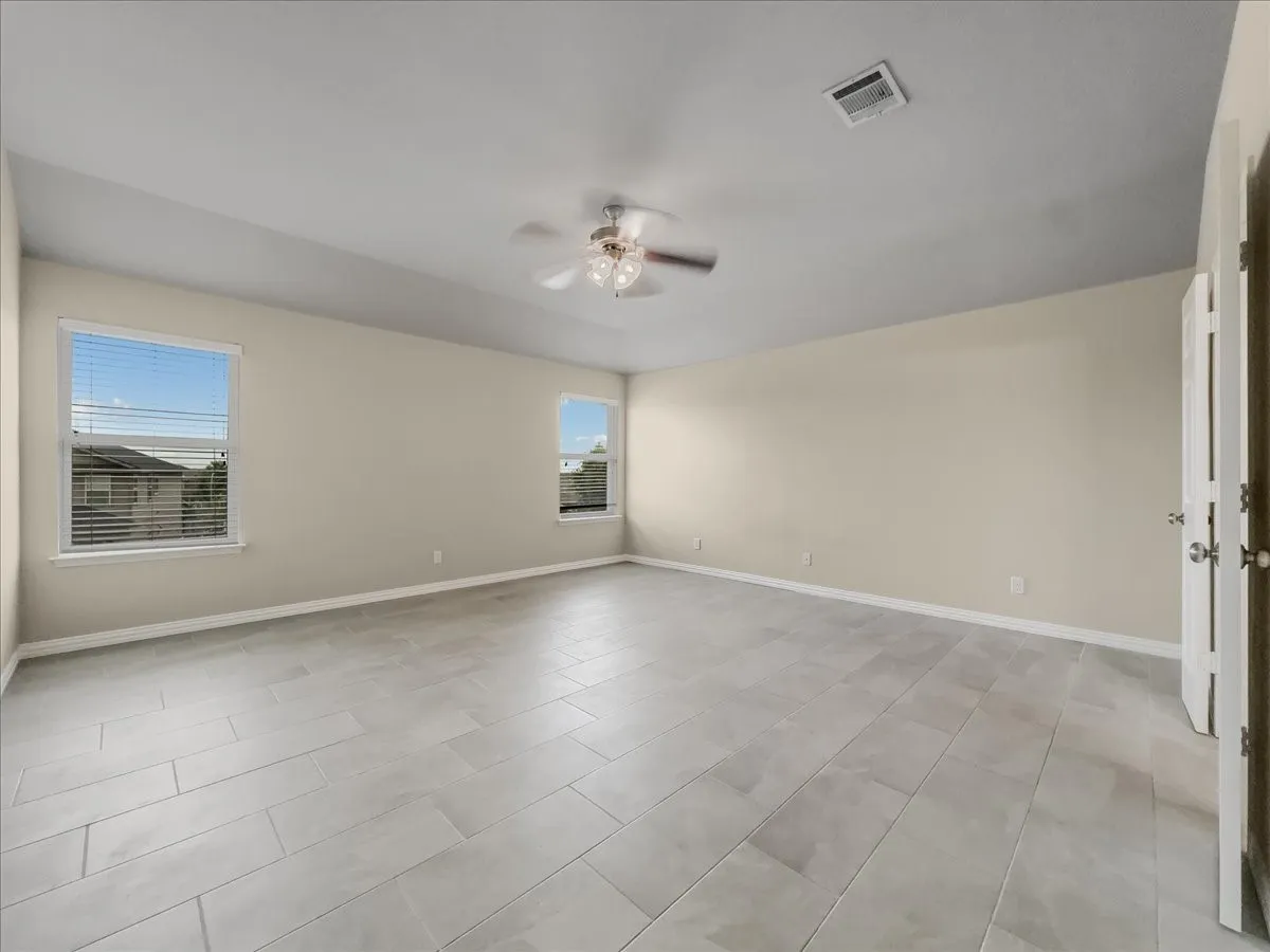 Spacious room featuring light-toned tile flooring, neutral wall paint, two windows with blinds, and a ceiling fan with integrated lighting
