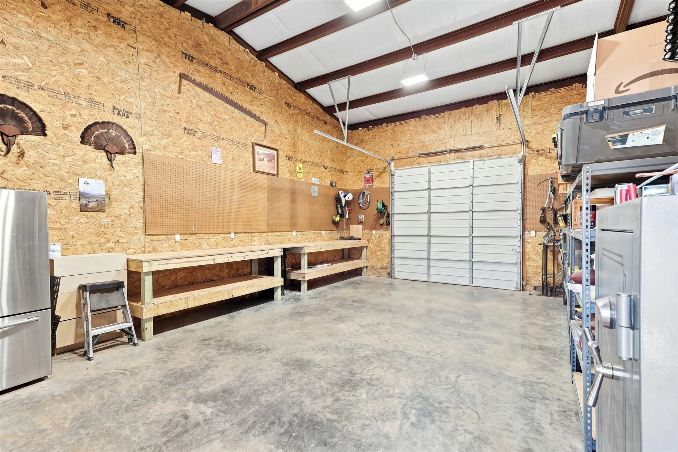 Spacious garage featuring exposed beam ceiling with skylights, cork composite wall paneling, polished concrete flooring, built-in workbench with storage shelving, and overhead garage door with opaque panels