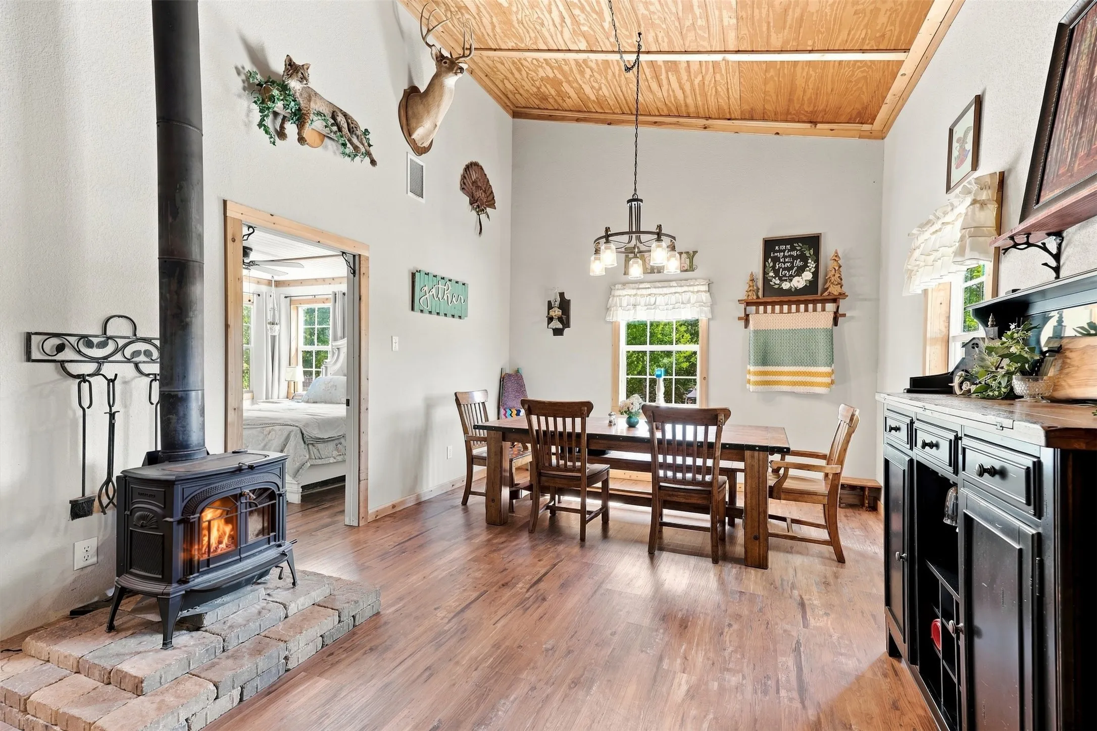Vaulted wood-paneled ceiling, wood-finish flooring, cast iron wood-burning stove on a brick hearth, and a chandelier with glass shades