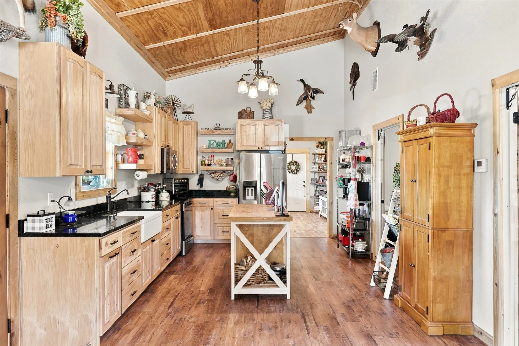Vaulted wood-paneled ceiling, wood-finish flooring, light wood cabinetry, black countertops, and a white farmhouse-style sink