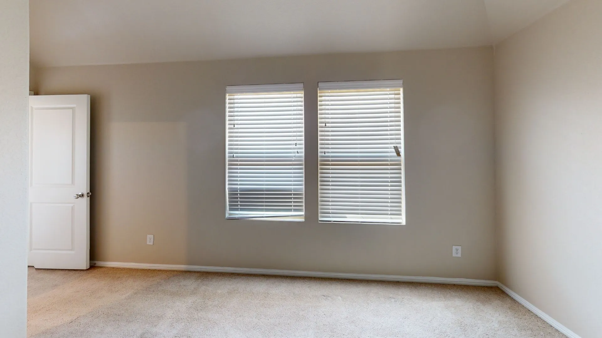 Neutral tone wall paint, carpet flooring, dual windows with blinds, and a white paneled door