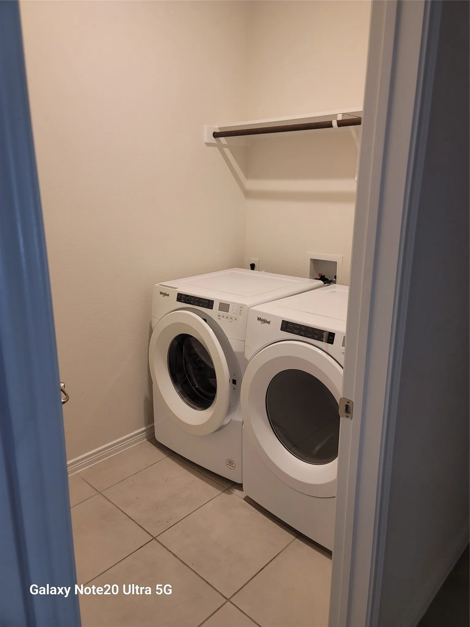 Laundry area with tile flooring, a white wall-mounted shelf, and a dark hanging rod