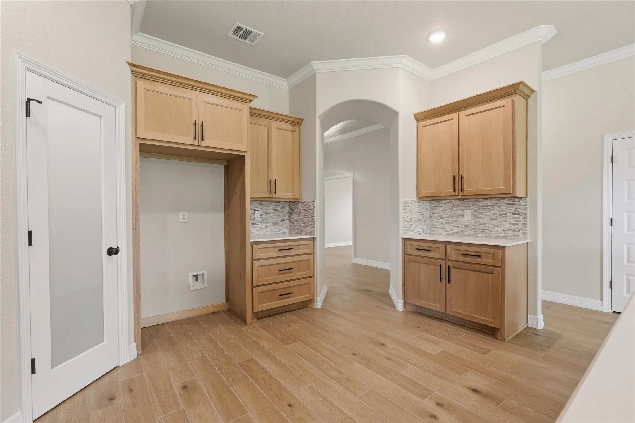 Wood-finish flooring, light wood cabinetry, stone-look backsplash, white countertops, and an arched entryway