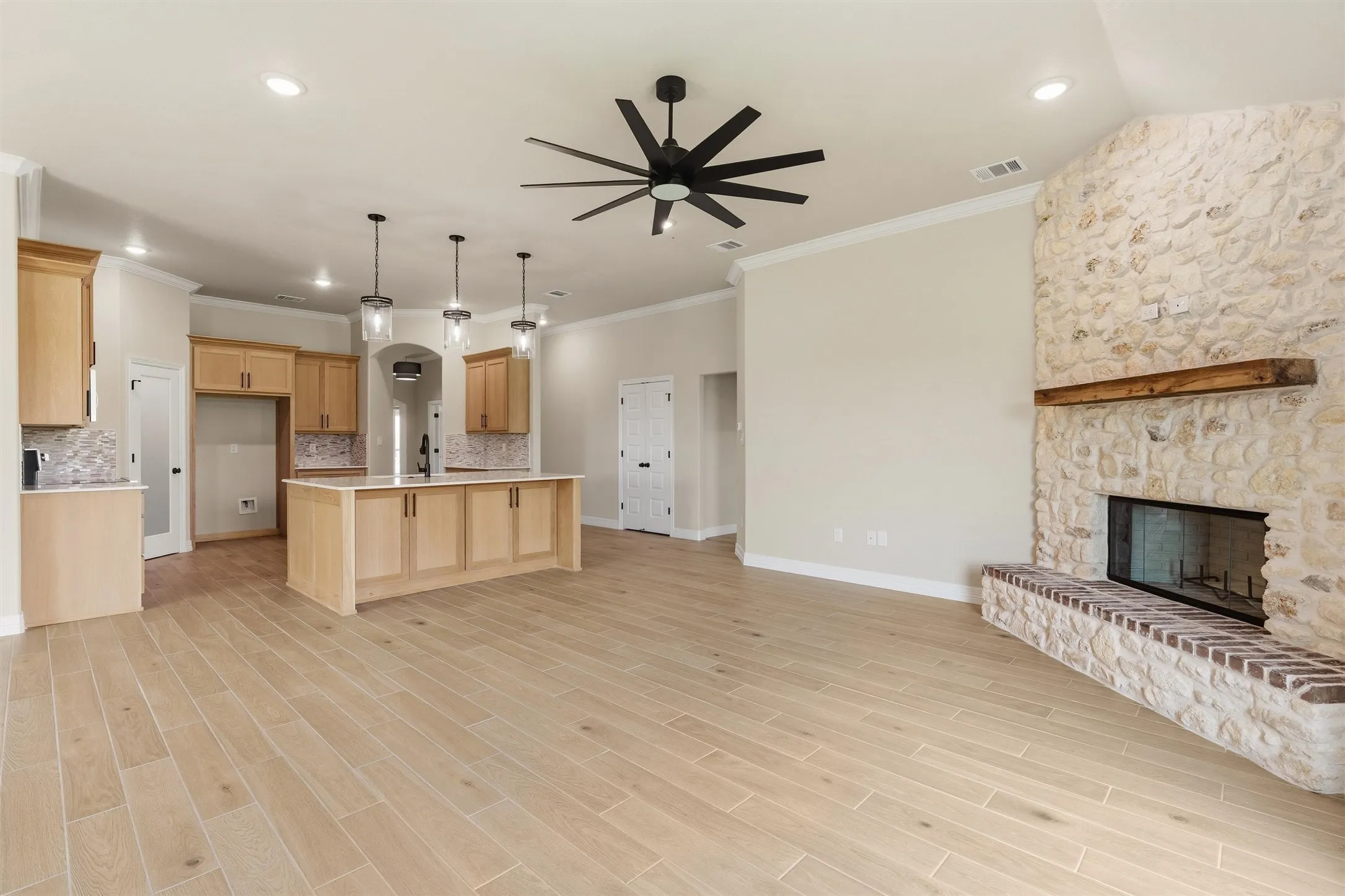 Open-concept living area featuring light wood-finish flooring, a stone fireplace with a wood mantel, a modern ceiling fan, recessed lighting, and a kitchen island with pendant lights