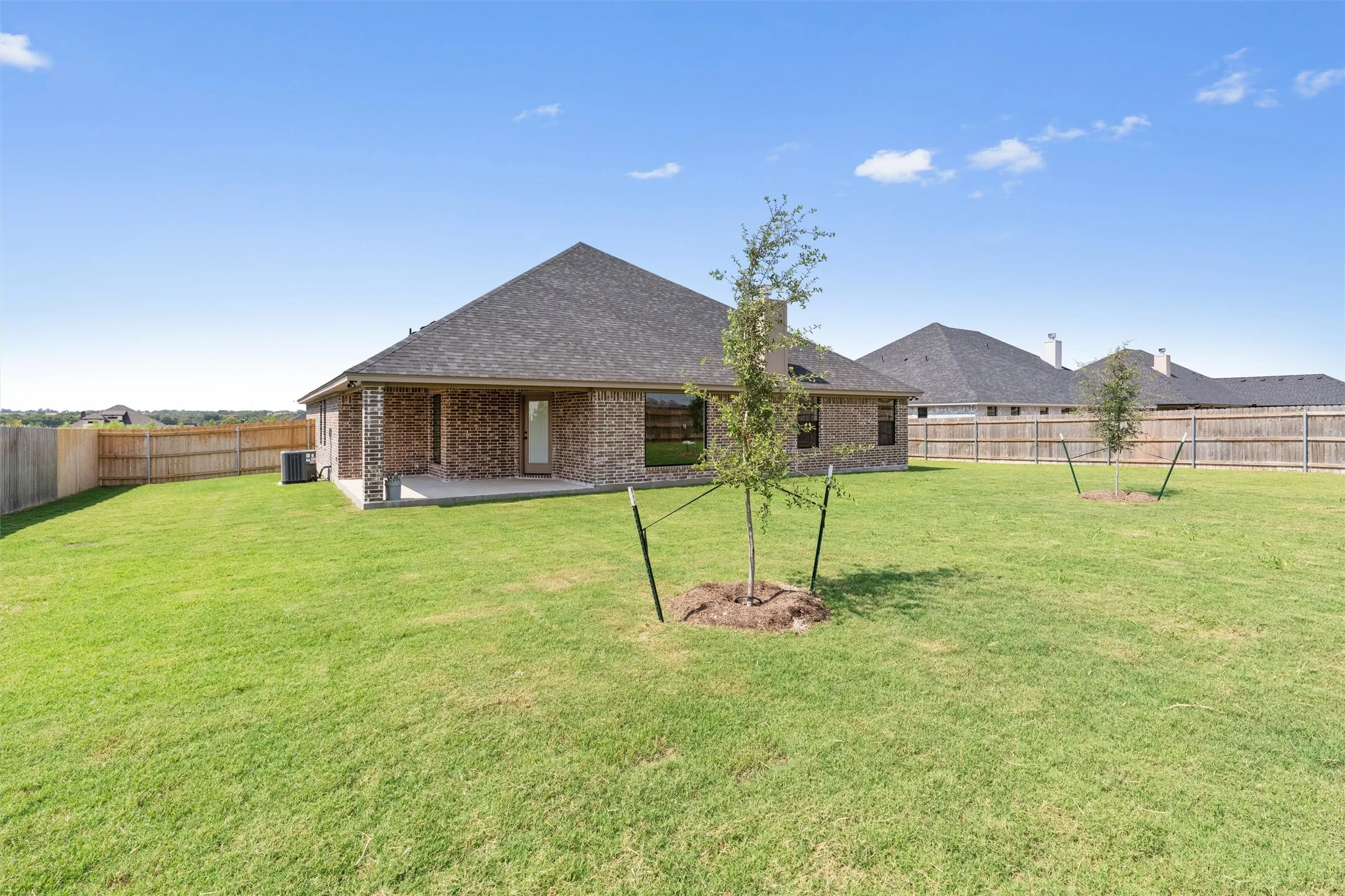 Expansive grassy yard with young trees, featuring a brick exterior with a covered patio, and a wood privacy fence