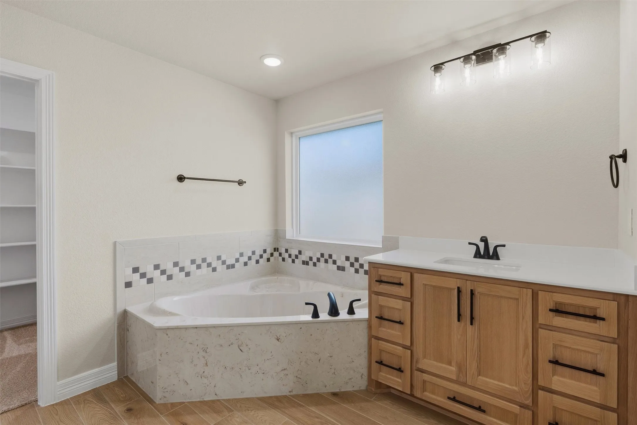 Corner soaking tub with tile surround, wood-finish flooring, single vanity with light wood cabinetry and white countertop, and a four-light vanity fixture