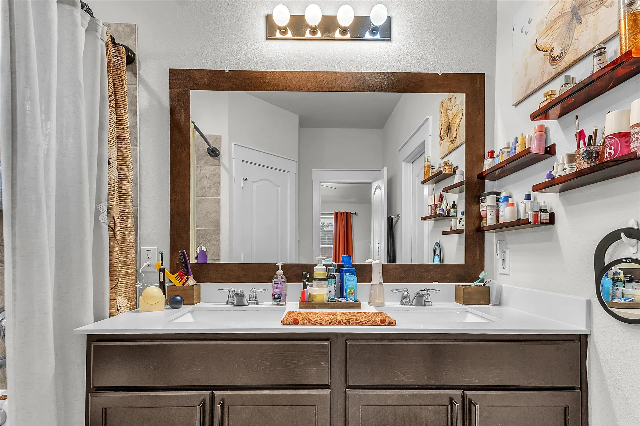 Bathroom with double vanity, quartz countertops, wood-framed mirror, wall-mounted shelves, and soft lighting from overhead fixture.