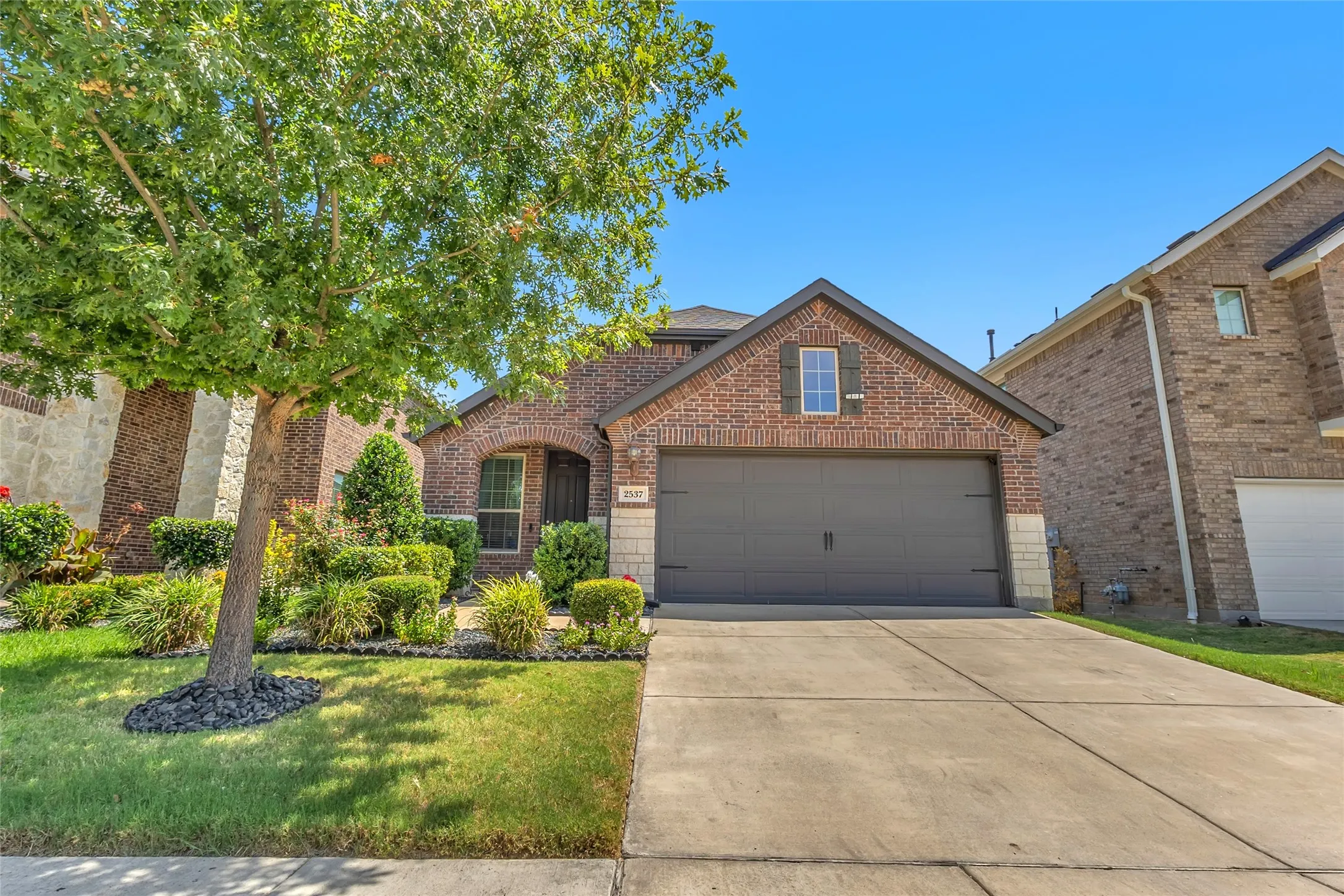 Single-story brick exterior with landscaped front yard, two-car garage, and concrete driveway under clear blue sky