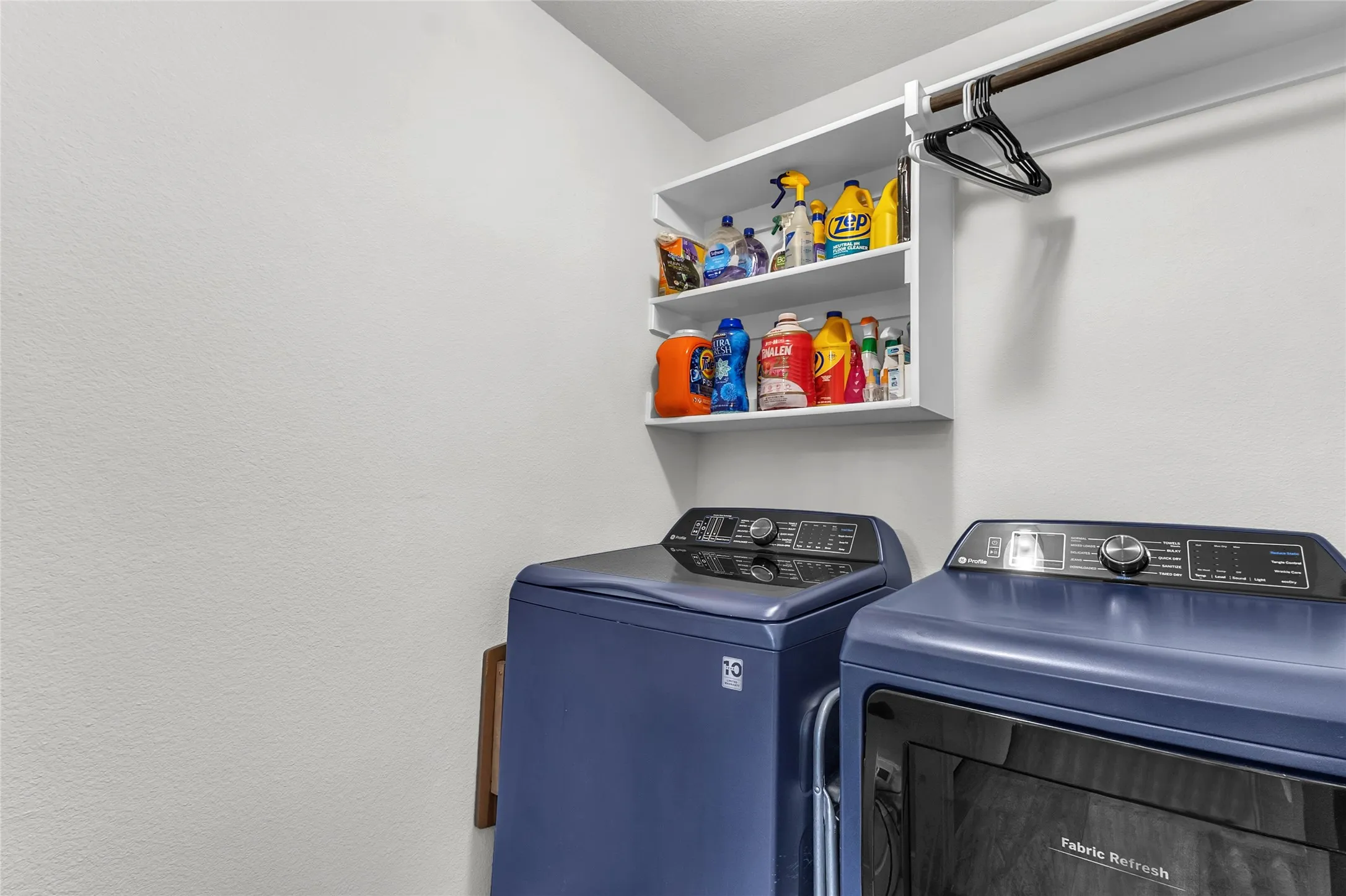 Laundry room with stacked blue washer and dryer, white shelving displaying various cleaning supplies, and neutral walls