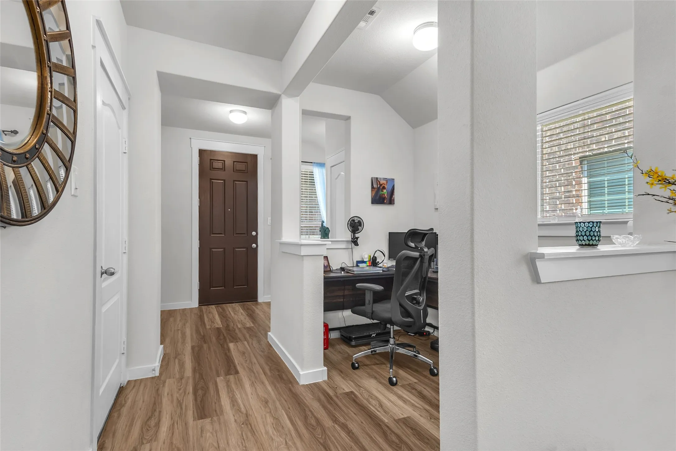 Entry hallway with hardwood flooring, white walls, and a dark wood front door, open layout leading to a study area with a desk and chair.
