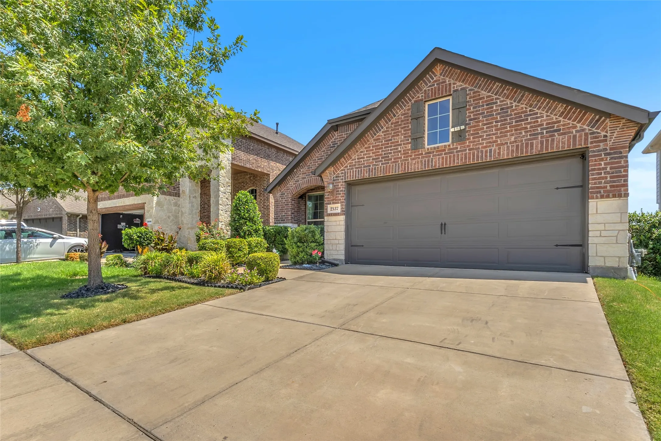 Two-car garage with a raised panel door, brick exterior, and landscaped front yard with a concrete driveway and mature trees.