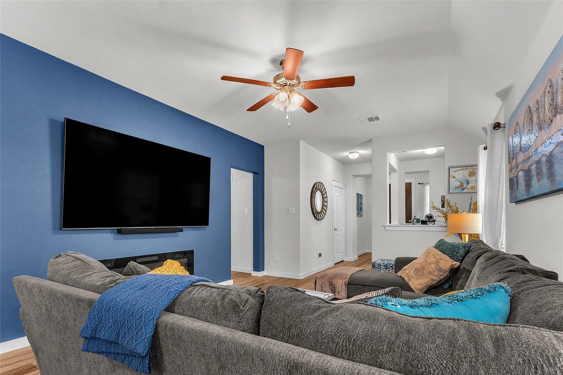 Living room with a large flat-screen TV, gray sectional sofa, hardwood flooring, blue accent wall, ceiling fan, and an open layout allowing natural light.