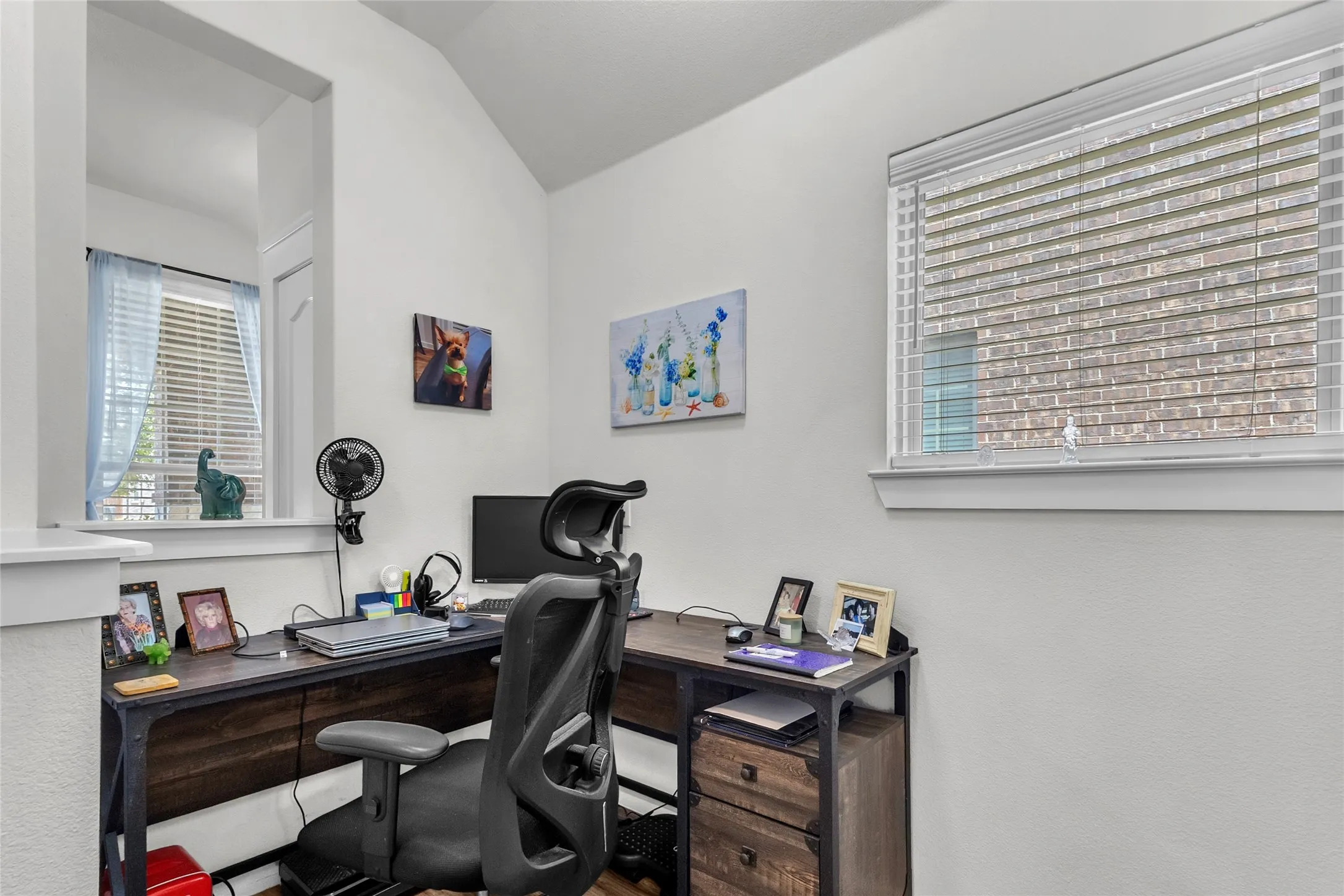 Home office with a wooden desk, computer setup, and natural light from the window, accented by framed photos and artwork on white walls.