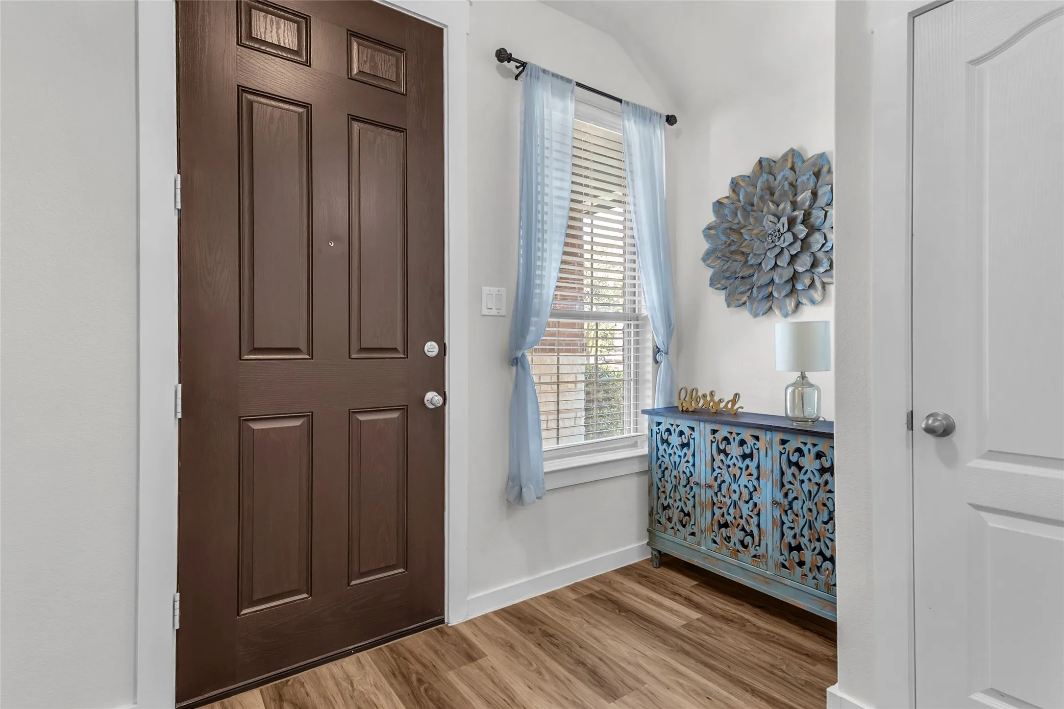 Entryway with dark wood door, light hardwood flooring, decorative blue console table, and sheer light-blue curtains over a window.