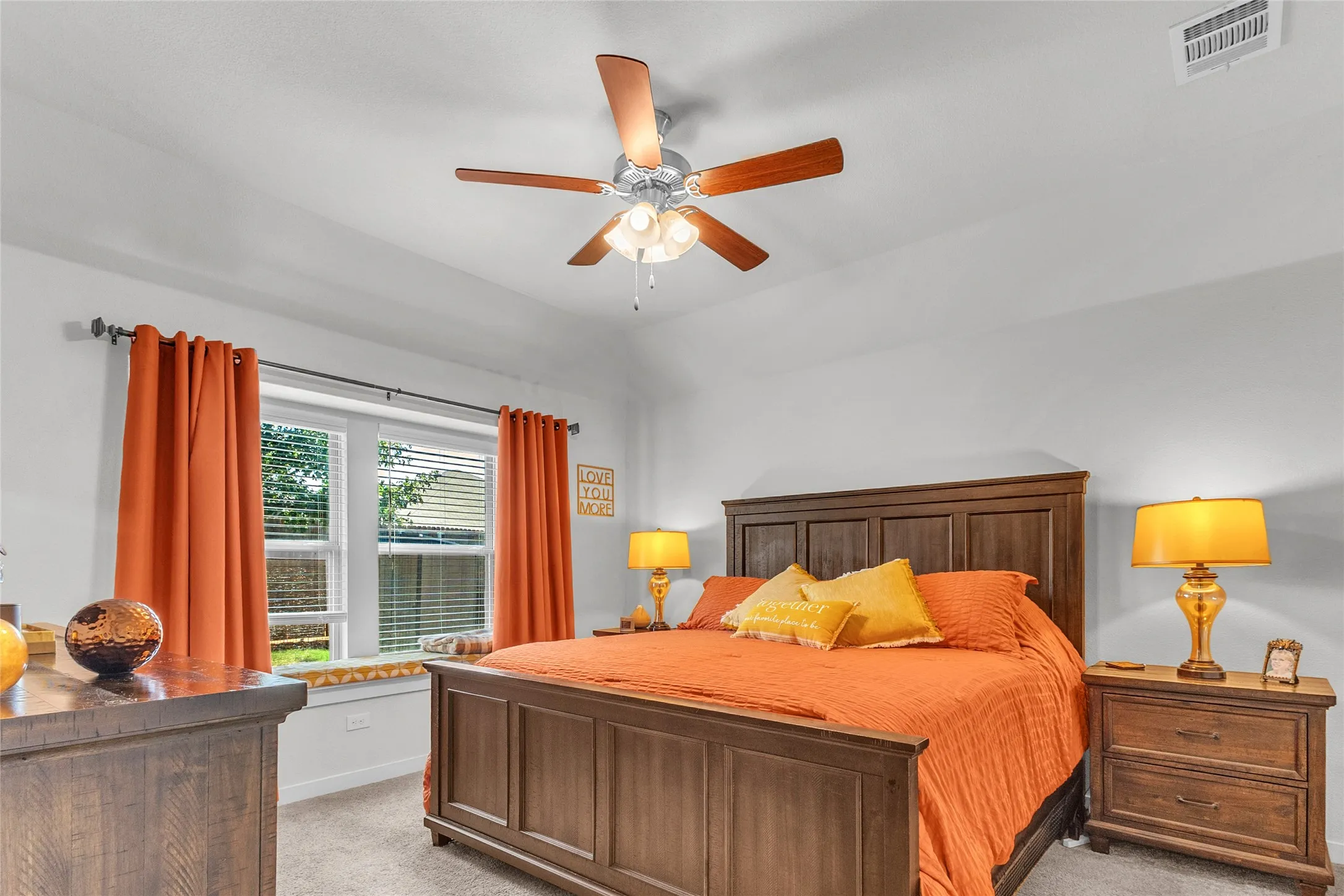 Primary bedroom with orange bedding, hardwood furniture, and ceiling fan, highlighted by natural light from large windows and soft carpet flooring