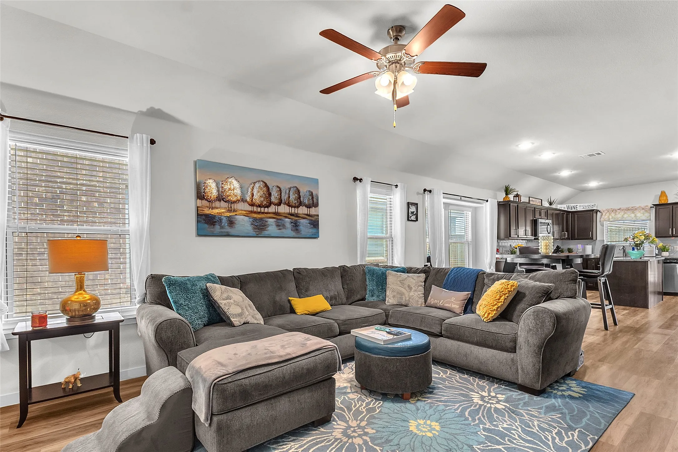 Living room with gray sectional sofa, ceiling fan, hardwood floors, decorative artwork, and an open-plan layout connecting to the kitchen area