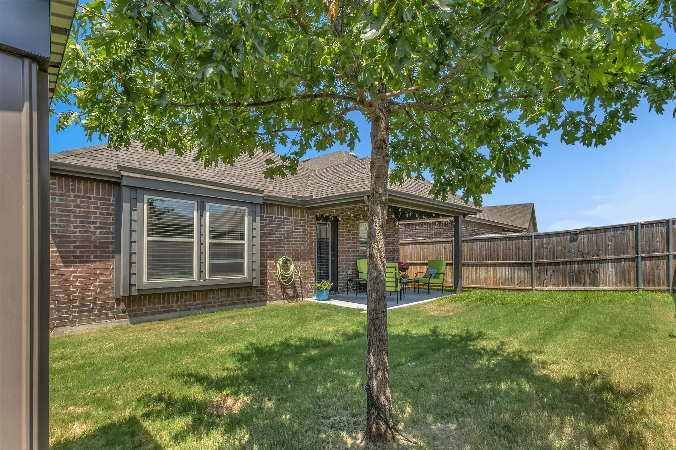 Covered back patio with a concrete surface, green lawn, single shade tree, and a fenced yard with seating area and potted plants.